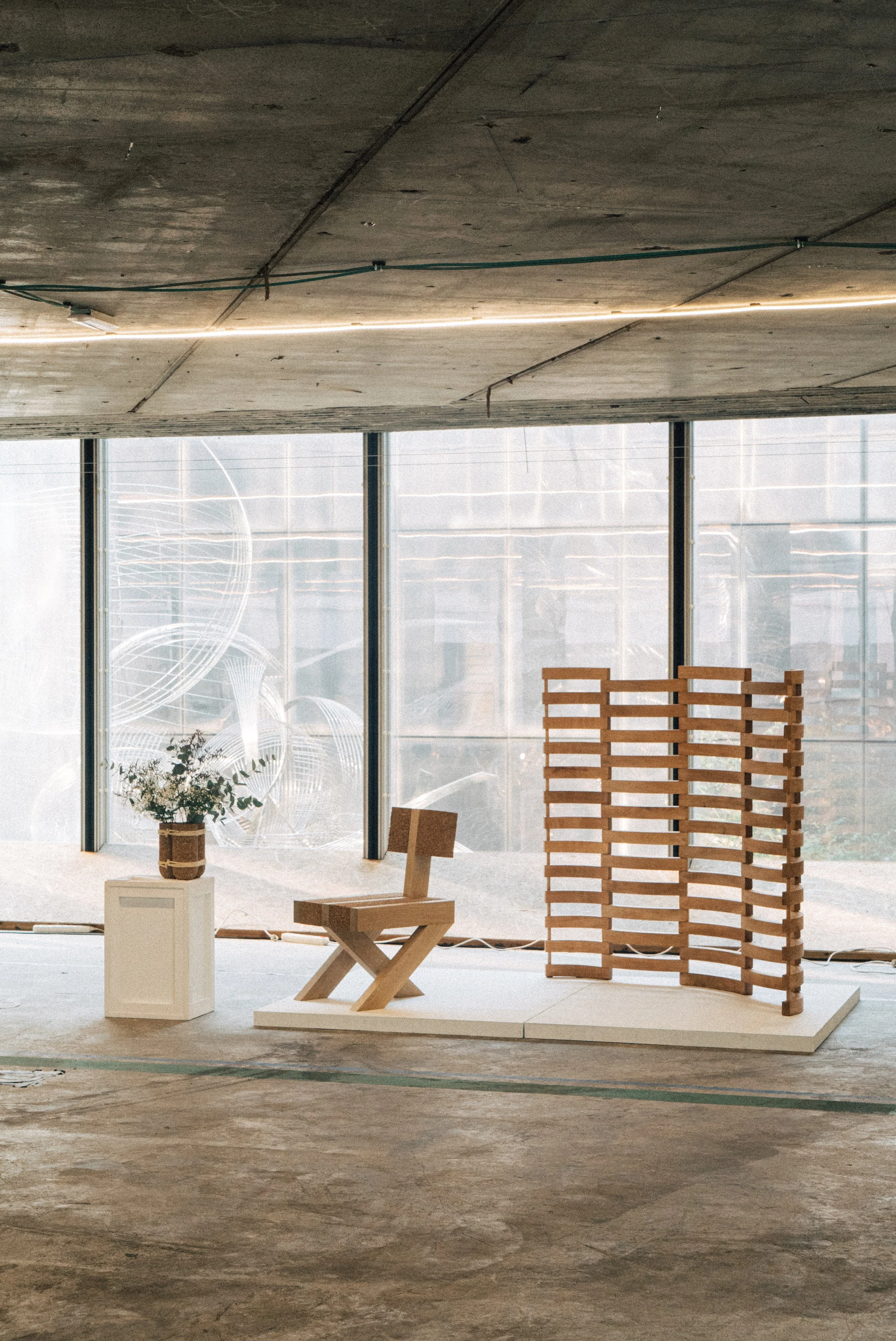 Interior space with a wooden chair, a small white pedestal with a potted plant, and a wooden room divider in front of large windows with a cityscape background.