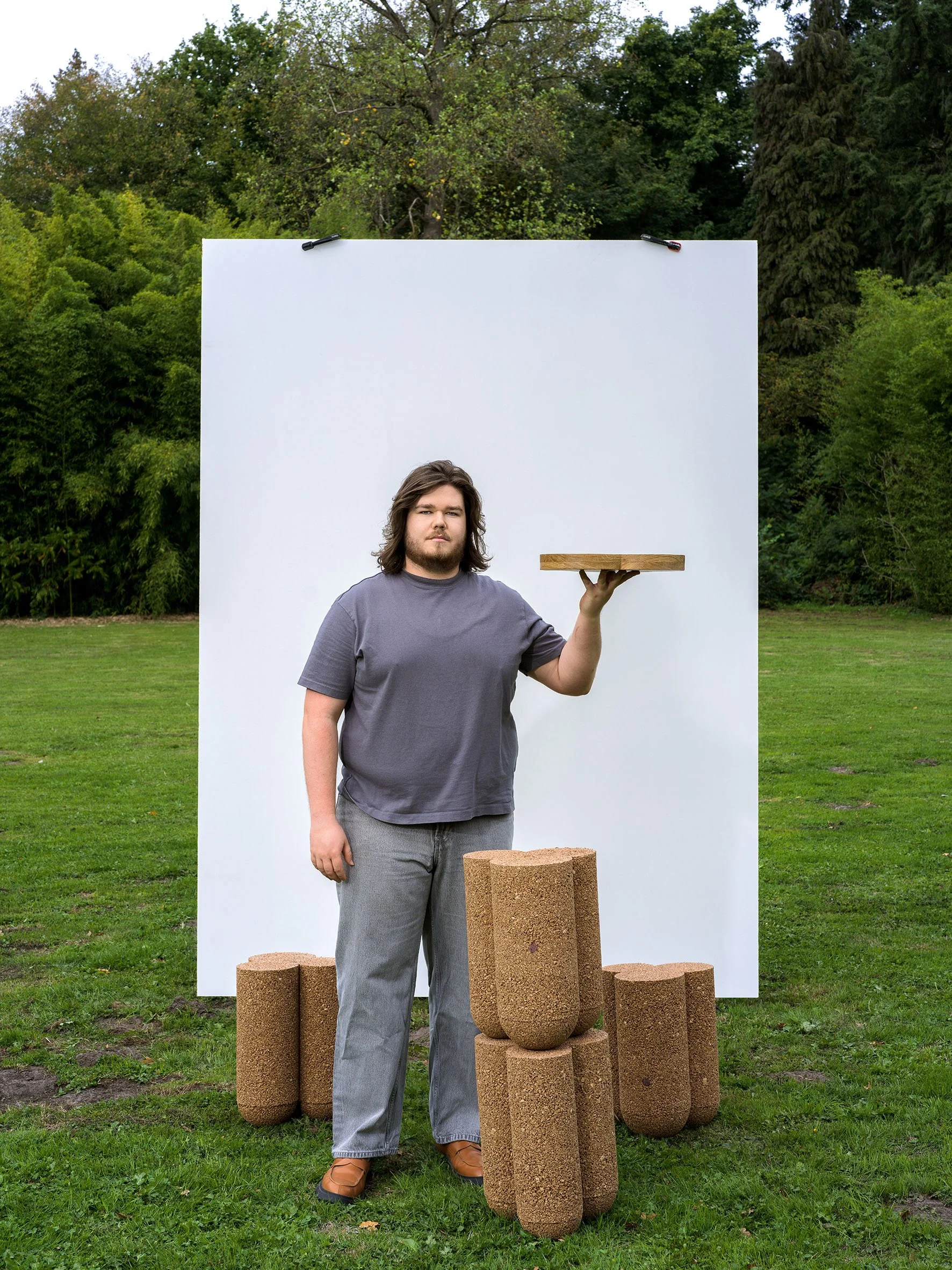 A man holding a wooden tray stands outdoors in front of a white backdrop, surrounded by large cork-like cylinders on grass.