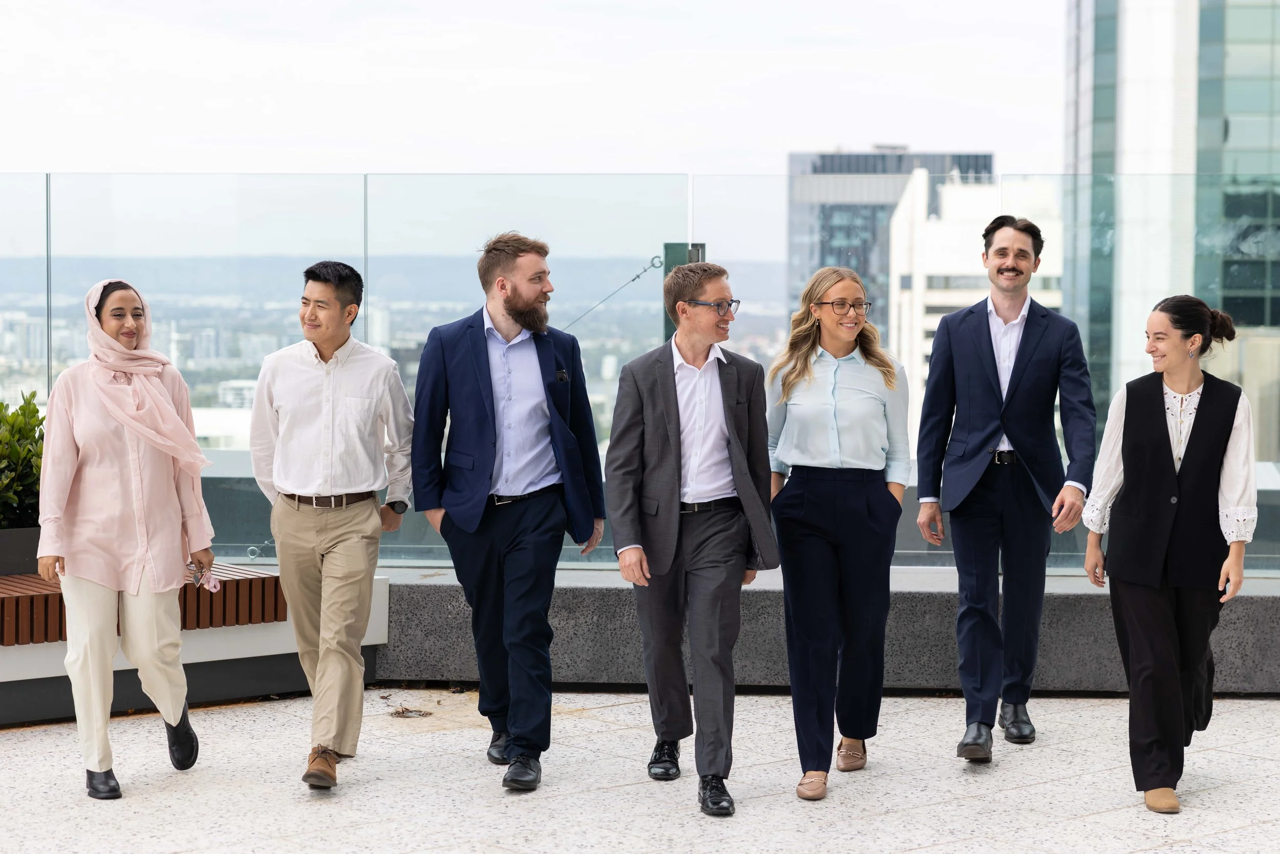 Group of seven diverse professionals walking and talking on a city rooftop balcony with glass railings and tall buildings in the background.