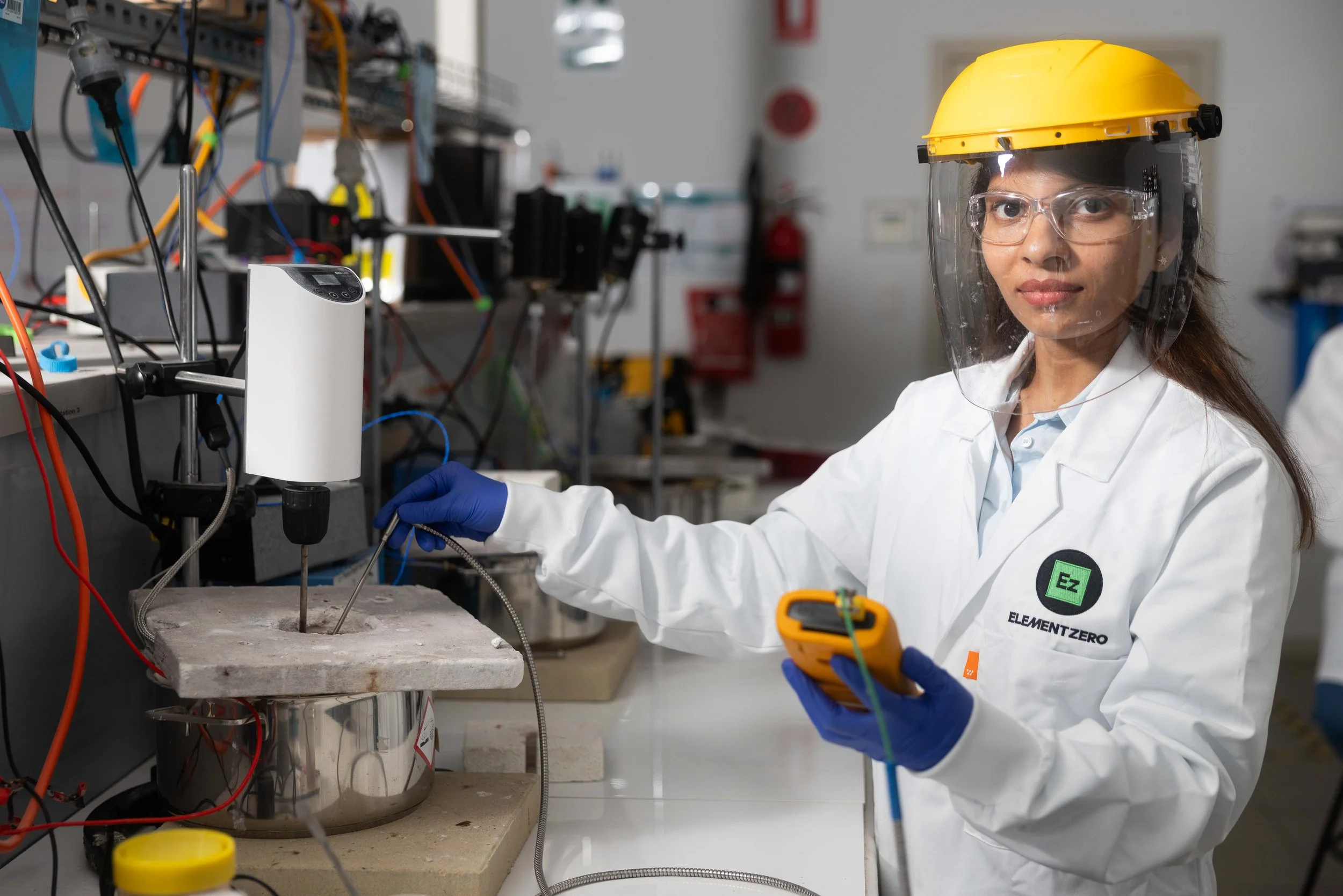 A female scientist wearing a yellow safety helmet, clear face shield, white lab coat with 'Element Zero' badge, and blue gloves working in a laboratory with scientific equipment and wires.