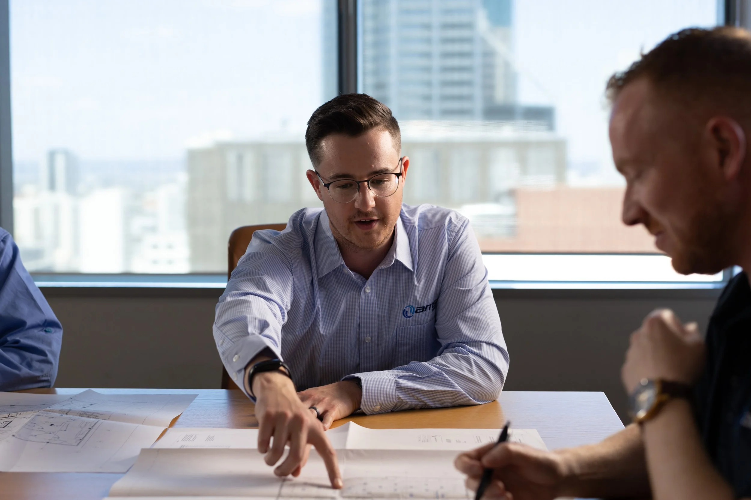 A man with glasses and a blue dress shirt sitting at a table in a modern office, explaining architectural plans to colleagues during a meeting.