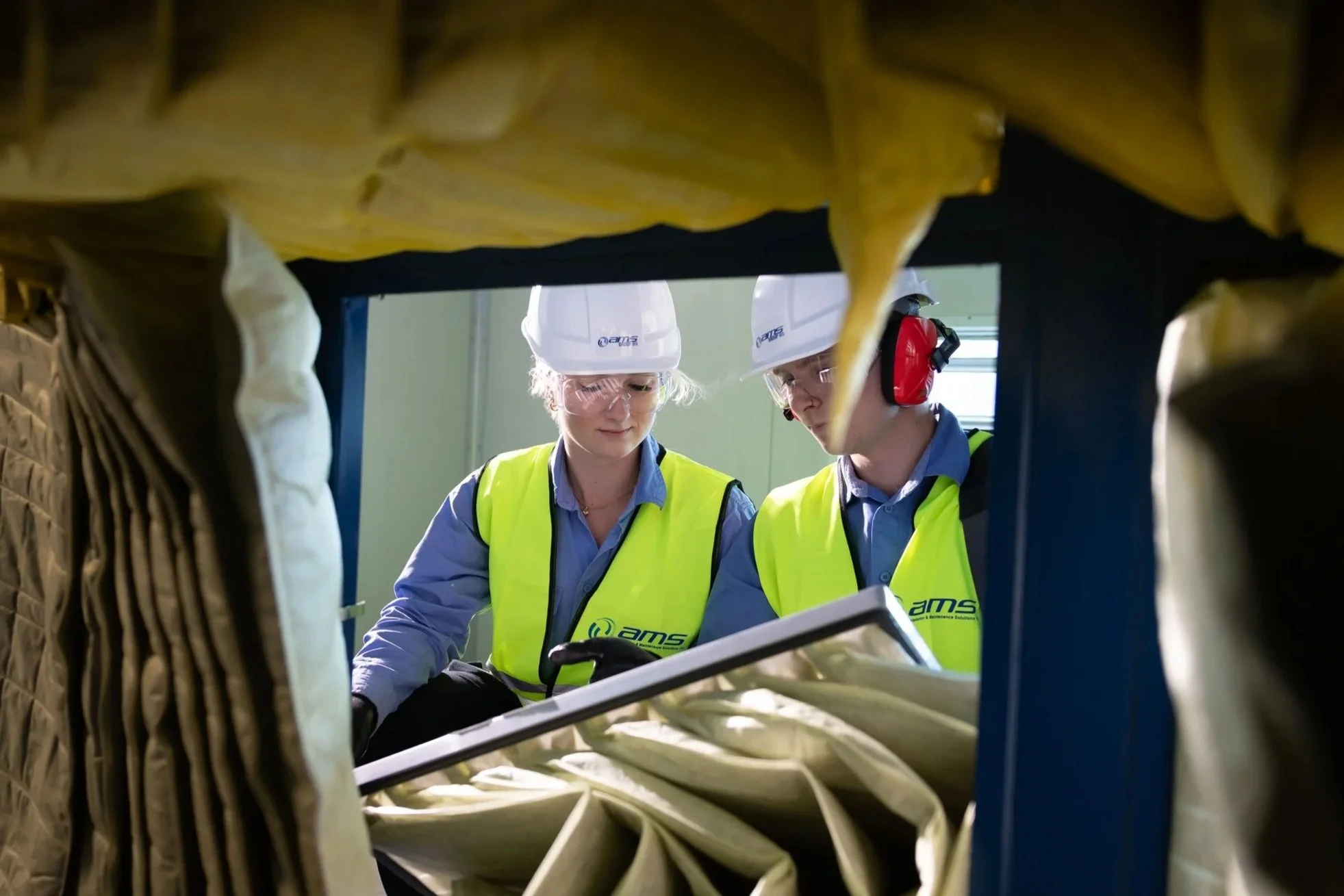 Two workers wearing helmets, safety glasses, and yellow vests inspecting equipment or machinery at an industrial site.