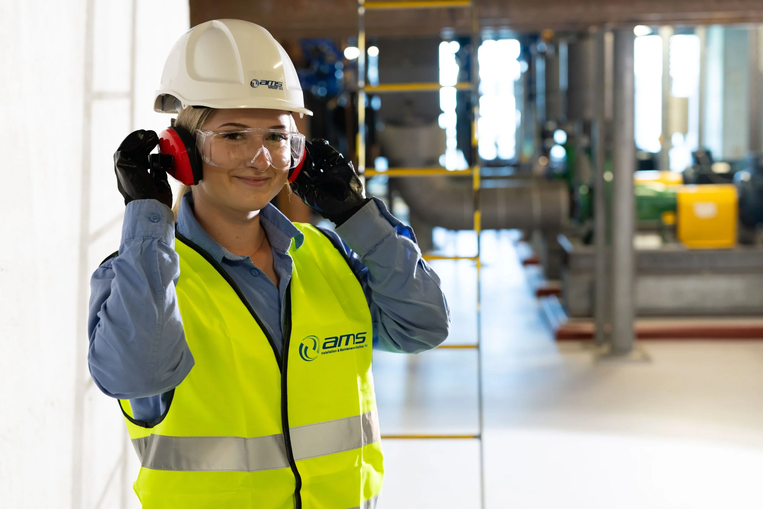 A woman in safety gear, including a white hard hat, clear safety glasses, red ear protection, black gloves, and a neon yellow safety vest, standing inside an industrial or construction setting with machinery and scaffolding in the background.