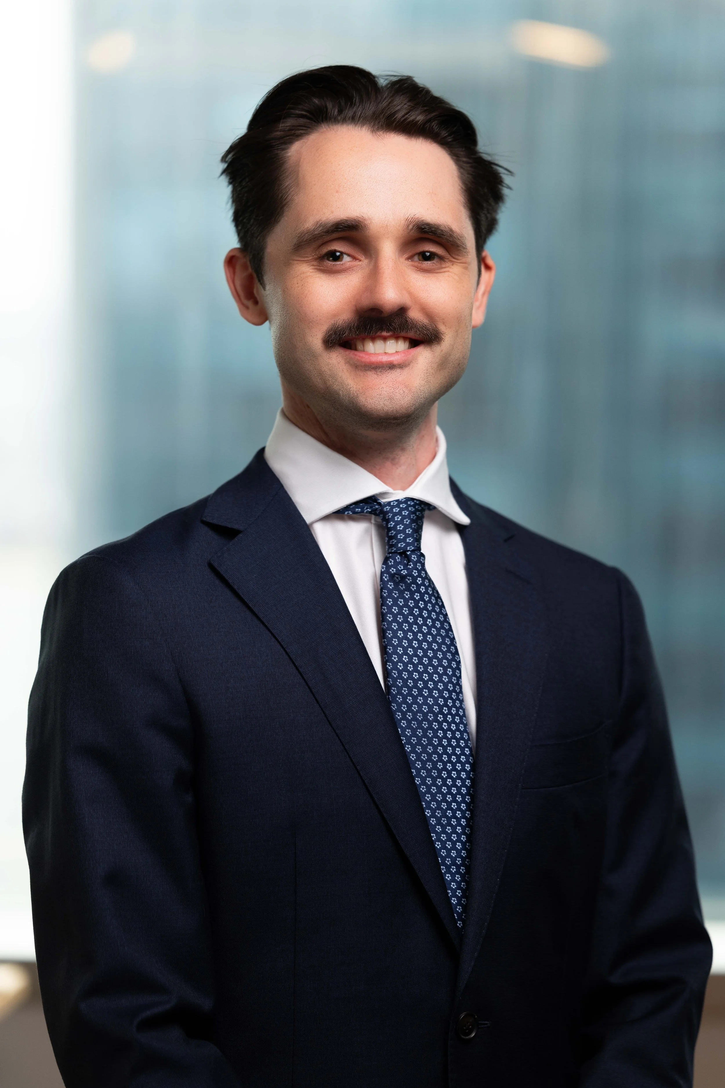 A professional man with dark hair, mustache, and a suit, smiling at the camera, in an office setting.