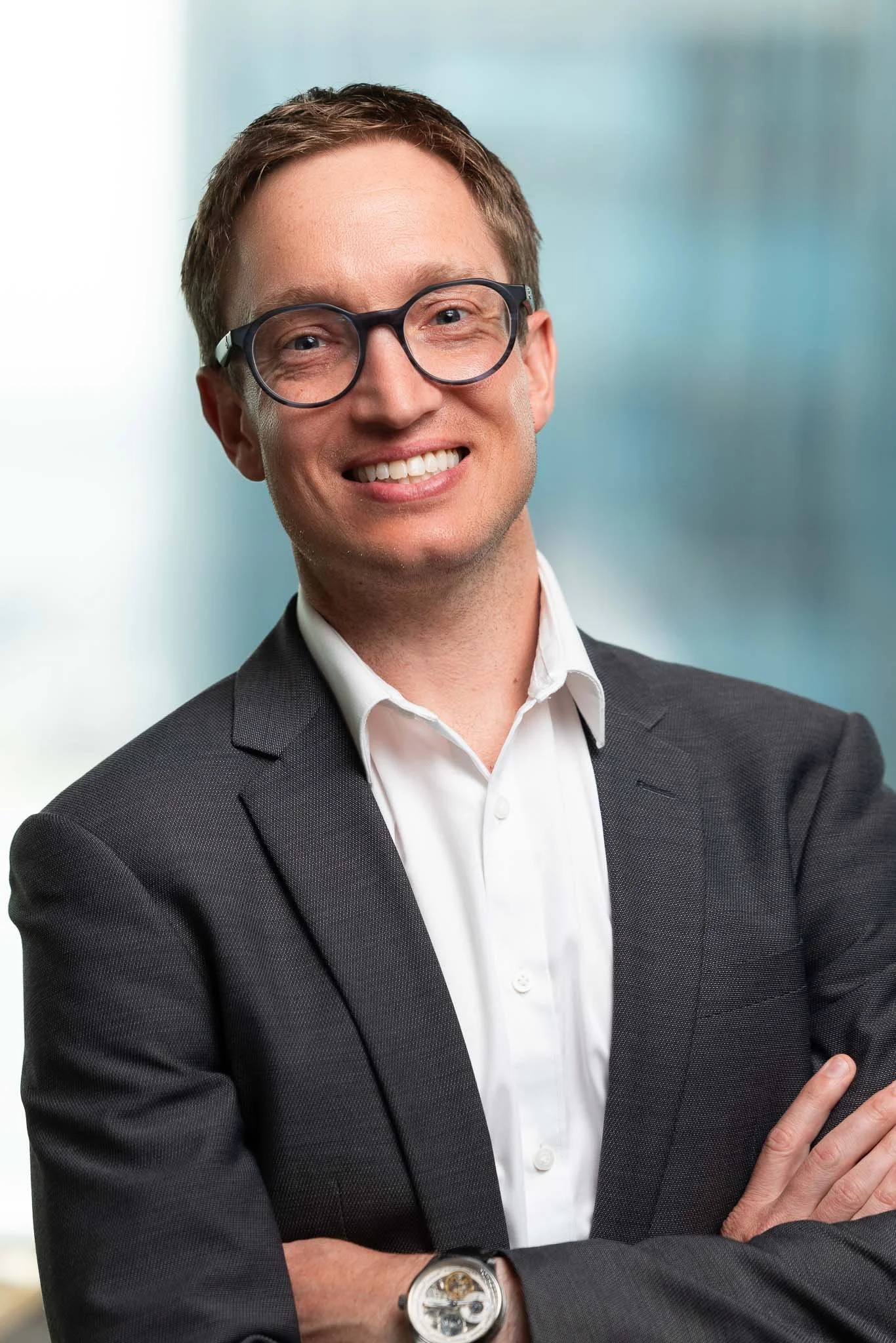 A smiling man with brown hair, wearing glasses, a dark gray suit, and a white shirt, standing with arms crossed in front of a blurred office background.