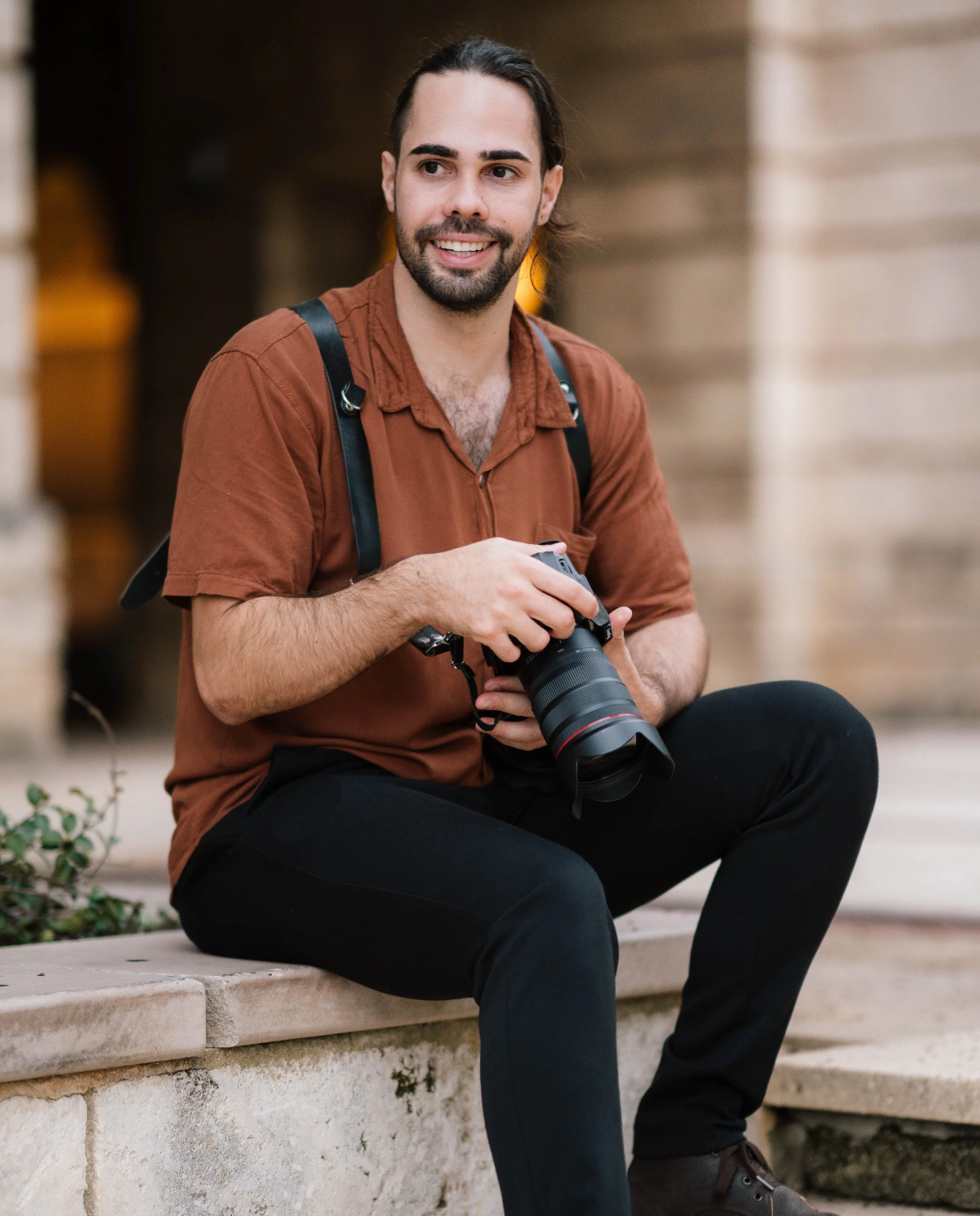 A young man with a camera sitting on a stone bench outdoors, smiling, wearing a brown shirt, black pants, and a black backpack.
