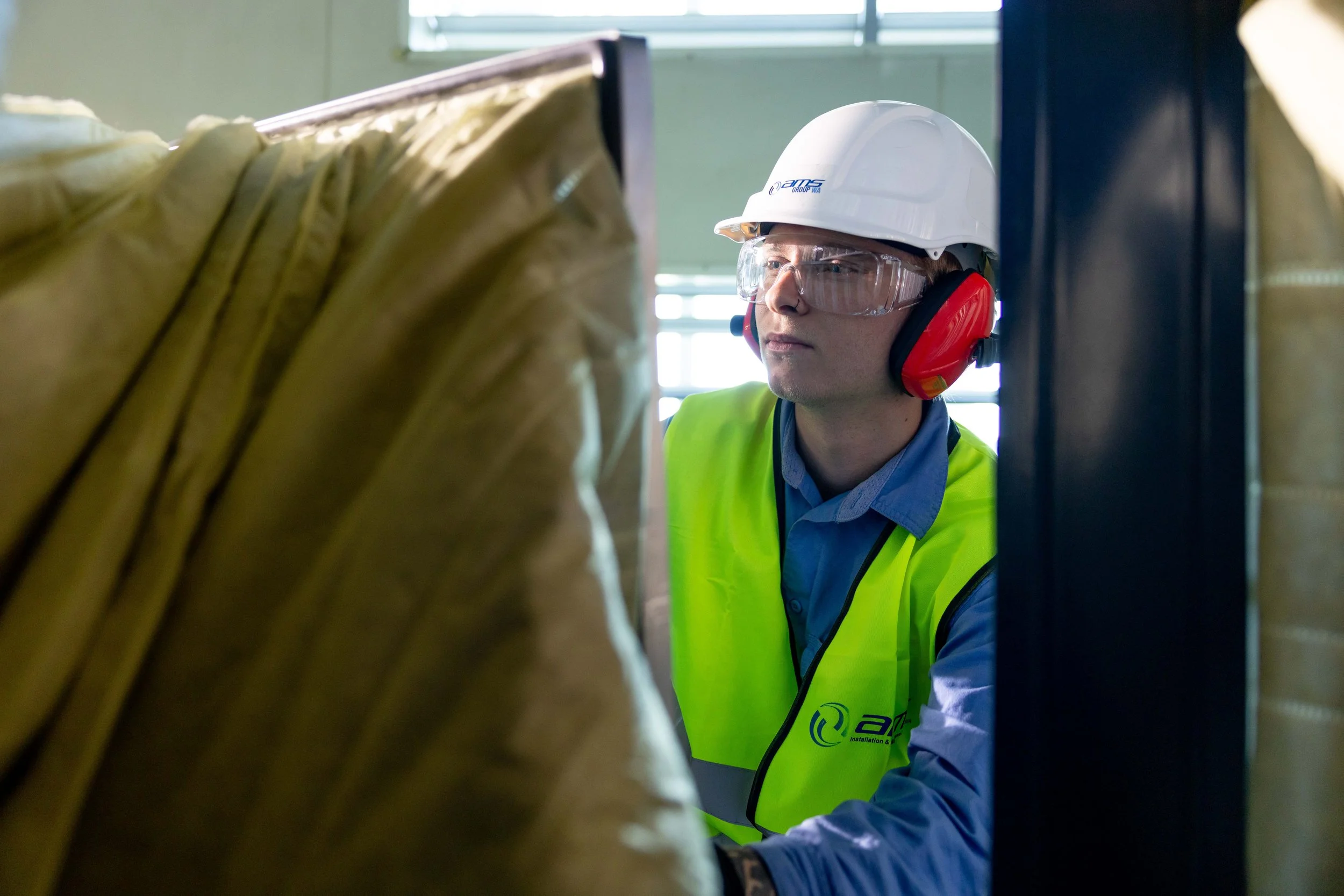 A male industrial worker wearing a white hard hat, clear safety glasses, red earmuffs, and a bright yellow safety vest looks at equipment in an industrial or warehouse setting.