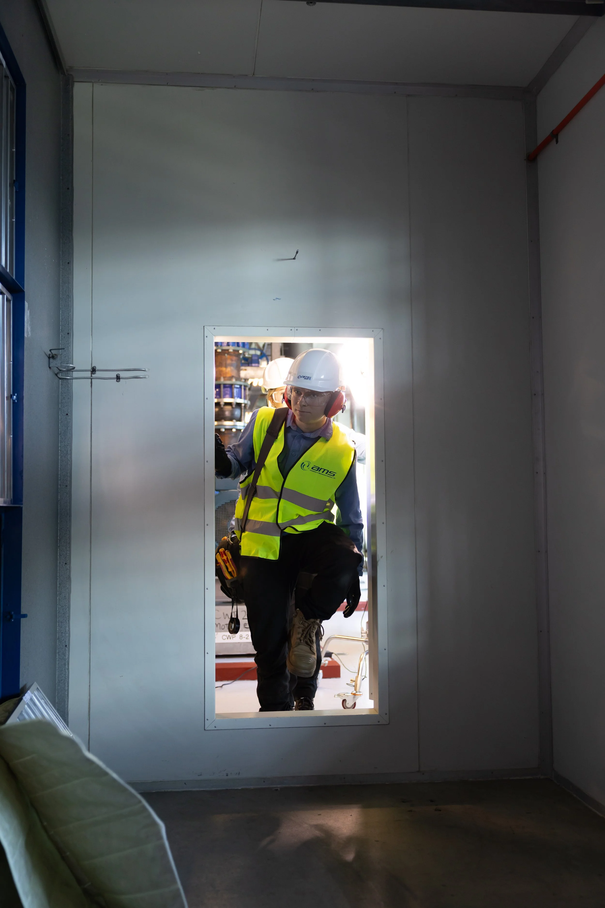 Construction worker wearing a white hard hat, safety glasses, a yellow safety vest, and gloves, kneeling at a doorway opening inside a construction site, with equipment and storage shelves visible in the background.