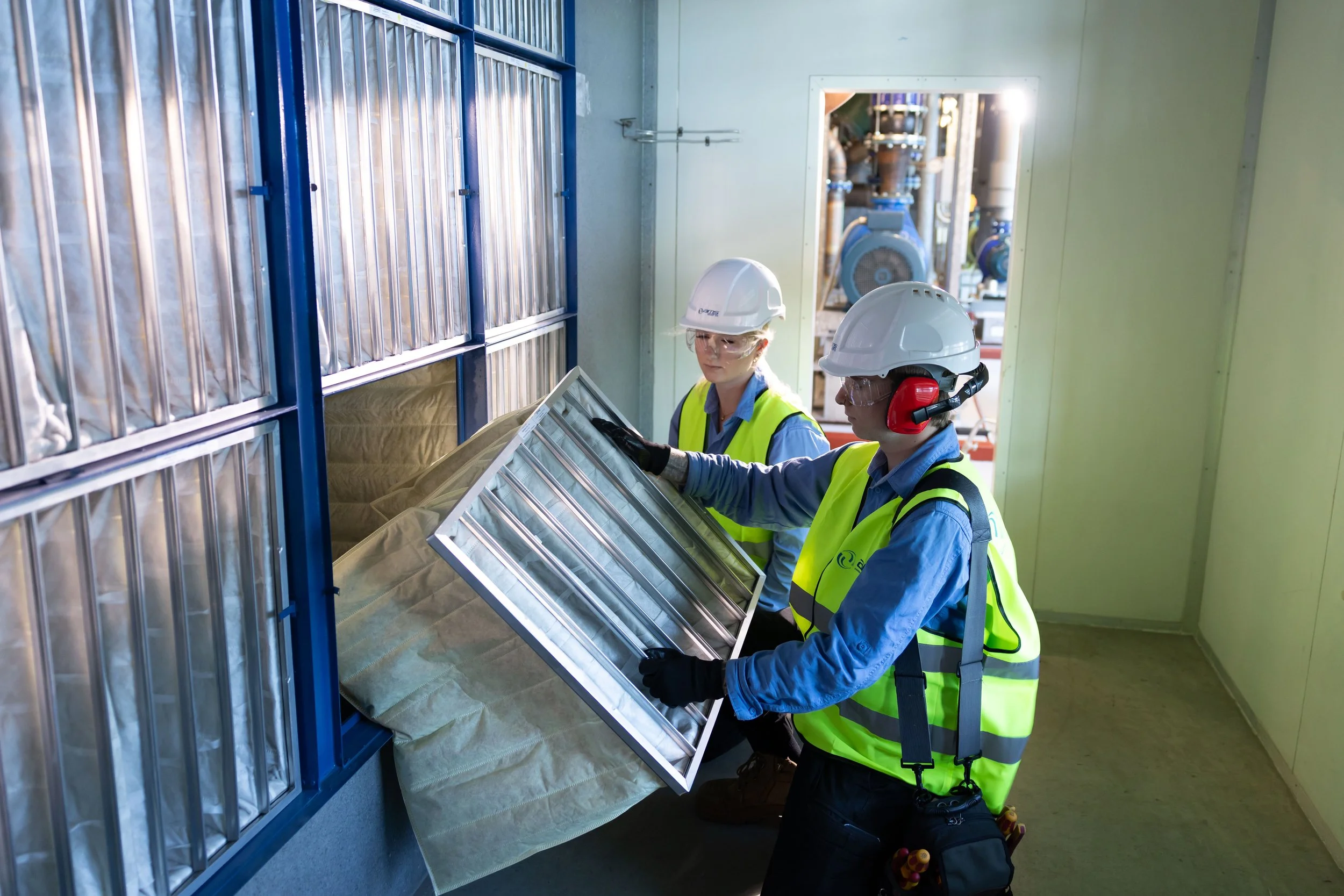 Two workers in safety gear inspecting equipment inside a facility. One is holding a large metallic panel, and there are blue and metallic structures around.