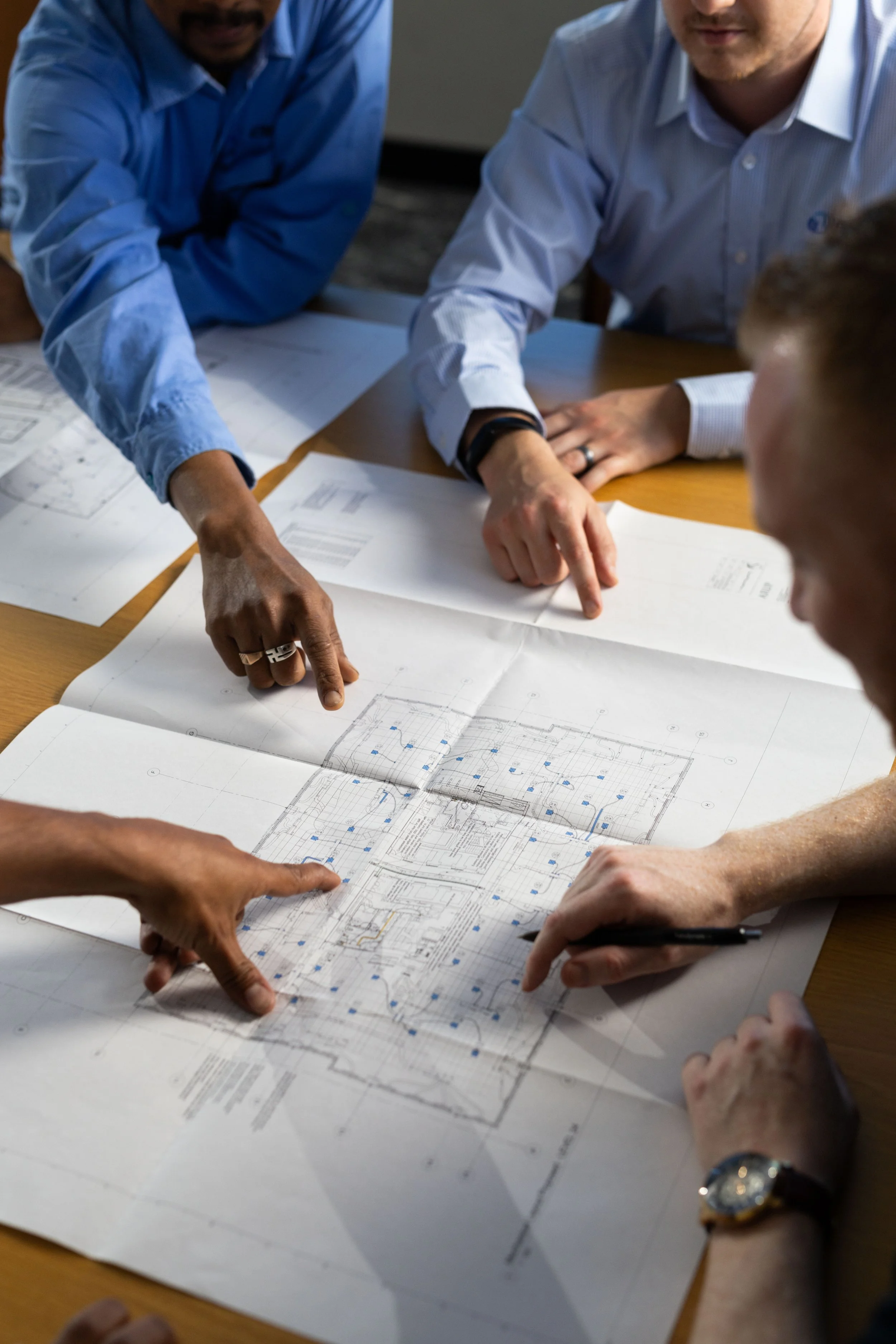 Group of people gathered around a table discussing blueprints or architectural plans, with hands pointing at different parts of the drawings.