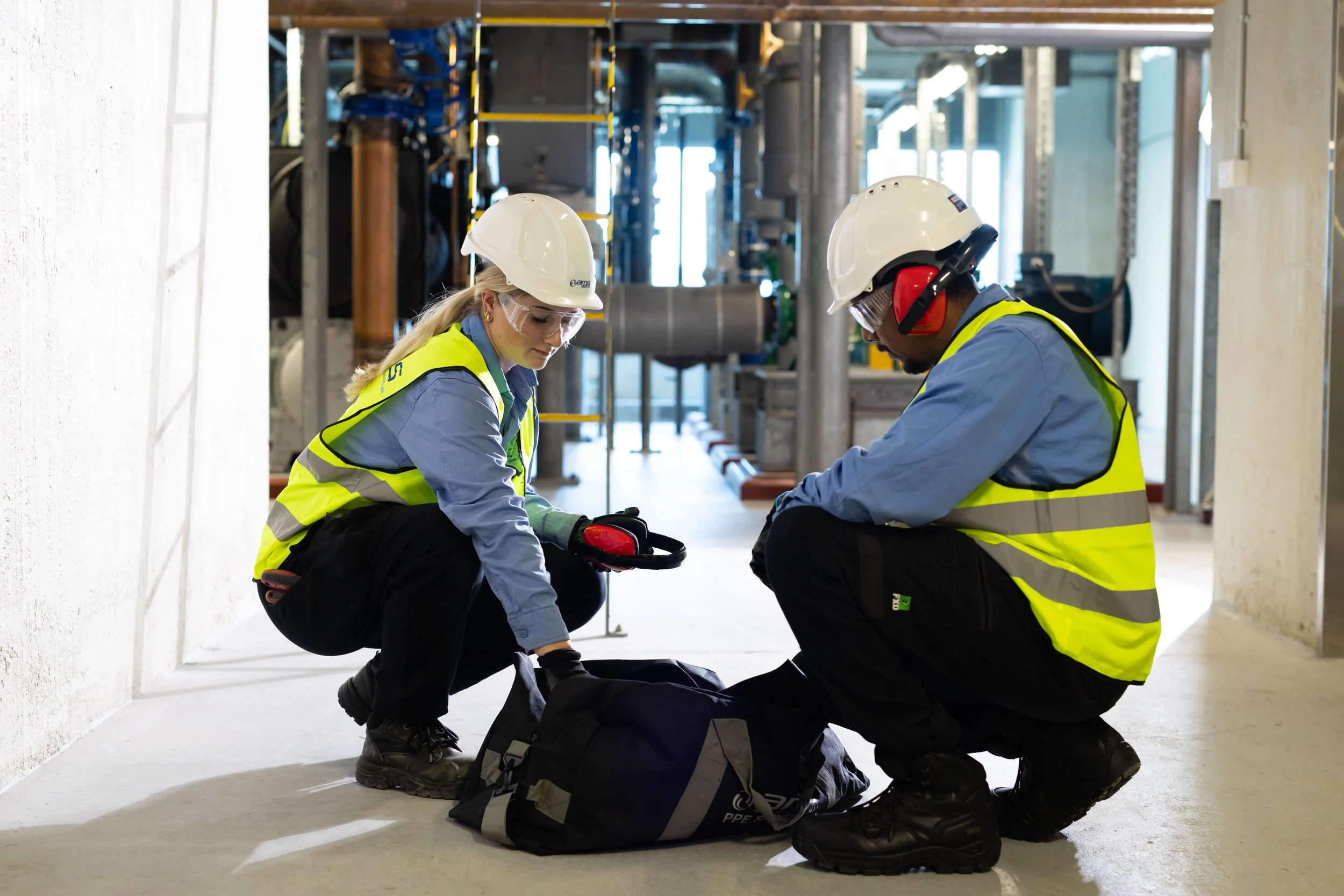 Two construction workers, a woman and a man, wearing high visibility vests, hard hats, and safety glasses, squatting on the floor and preparing equipment or tools at a construction site.