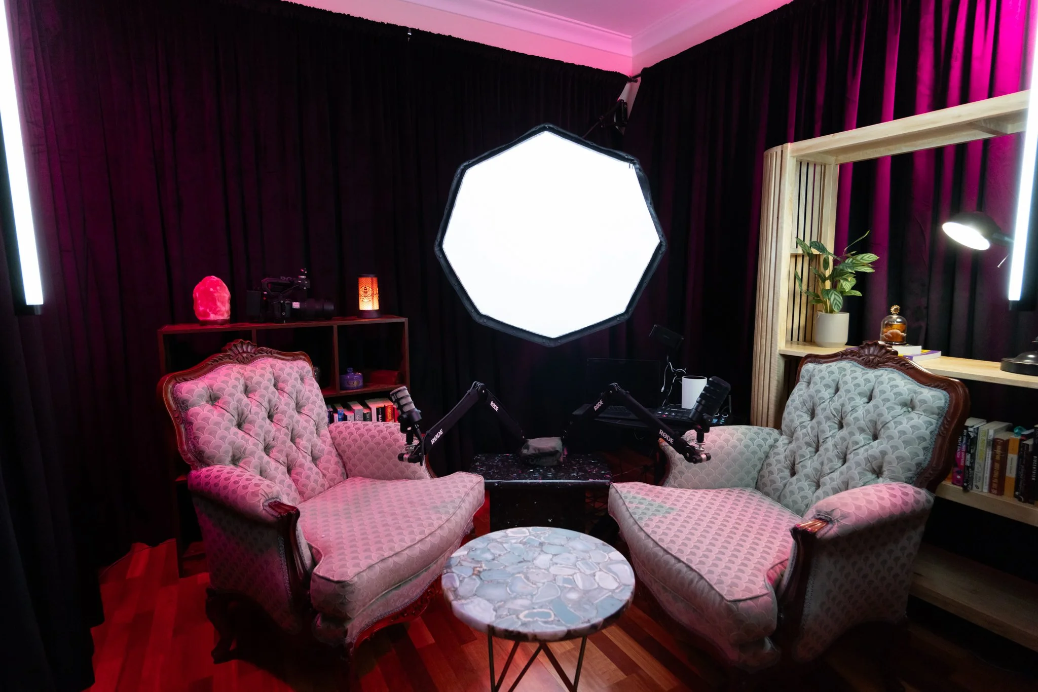 A recording or streaming setup with two vintage sofas, a circular marble coffee table, a bookshelf, a salt lamp, and two microphone arms, with professional lighting and a large hexagonal softbox centered on a dark wall.