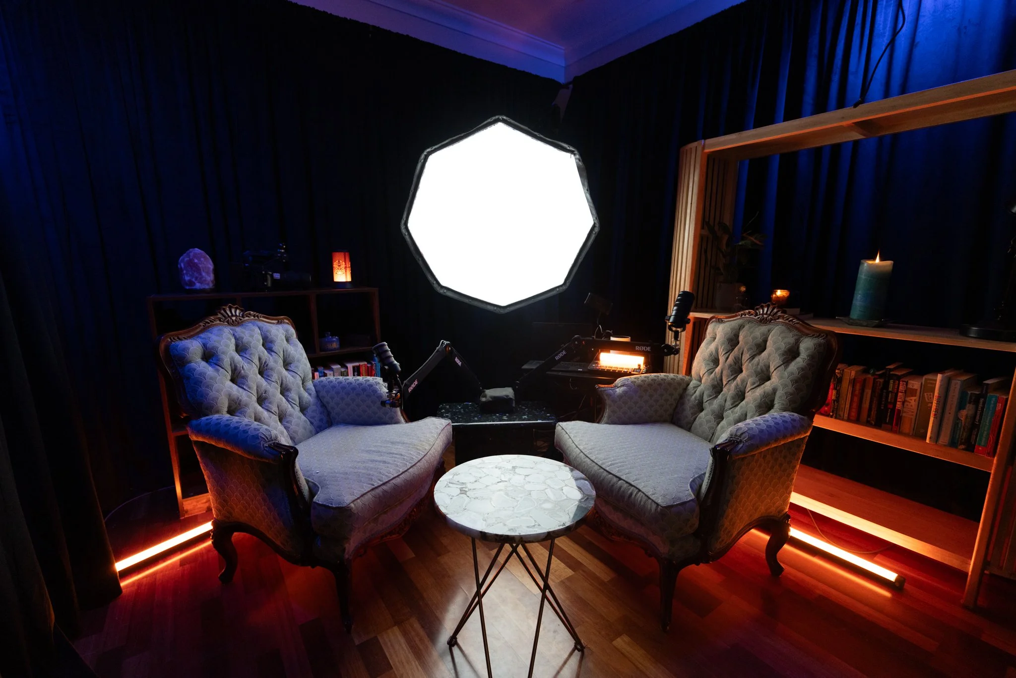 A recording or podcast studio setup with two vintage armchairs facing each other, a small round marble-topped table in between, a large bright octagonal light on the wall, a bookshelf with books, candles, and decorative items, dark curtains, and various recording equipment.