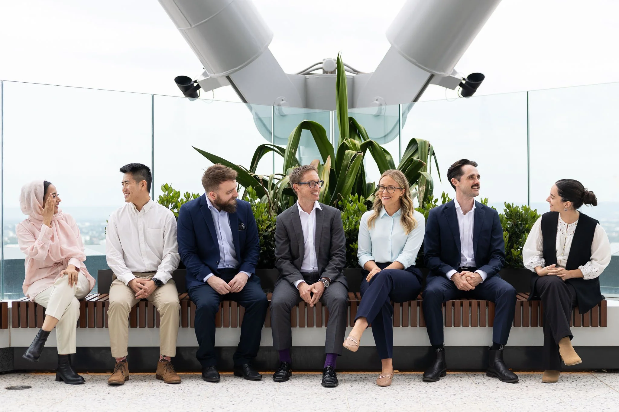 A diverse group of seven professionals sitting on a bench on a rooftop, engaged in conversation, with a large plant and glass railing in the background.