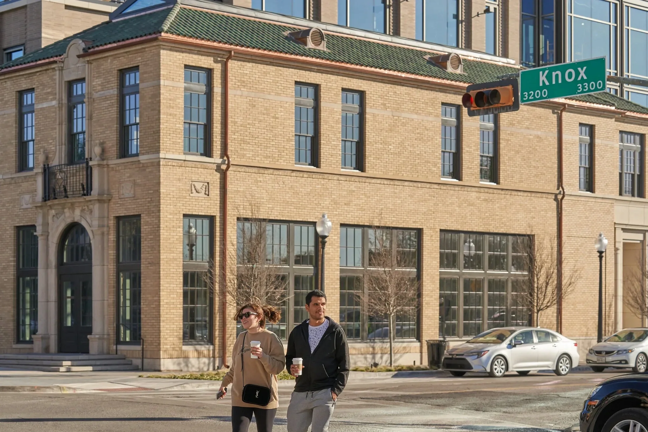 Two people walking across a city street with coffee cups, in front of a brick building with large windows, and a street sign that reads Knox.