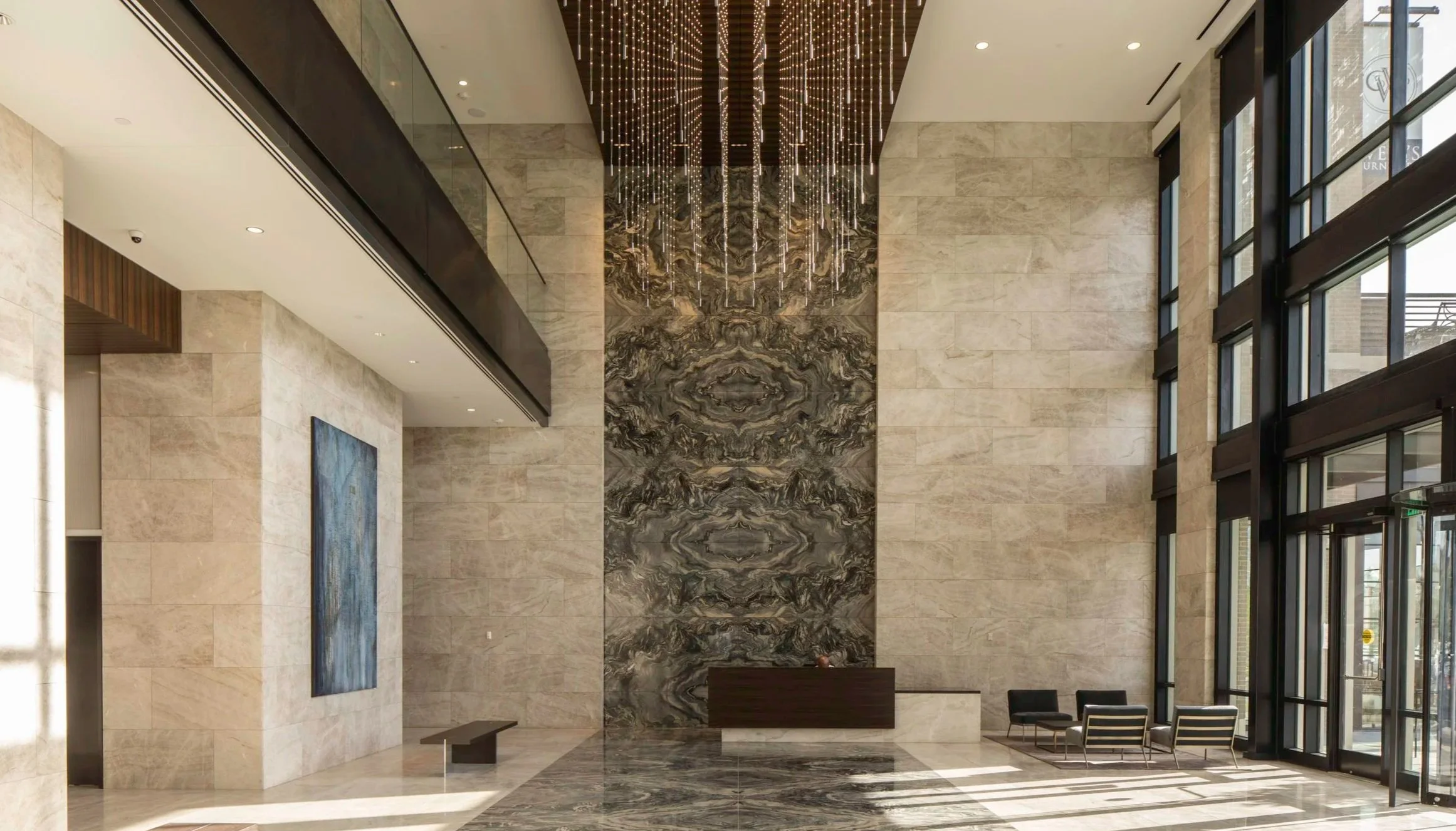 Modern hotel lobby with beige stone walls, a marble accent wall with a symmetrical pattern, and a glass entrance with sunlight streaming in. There are a few black lounge chairs and a dark wood reception desk.
