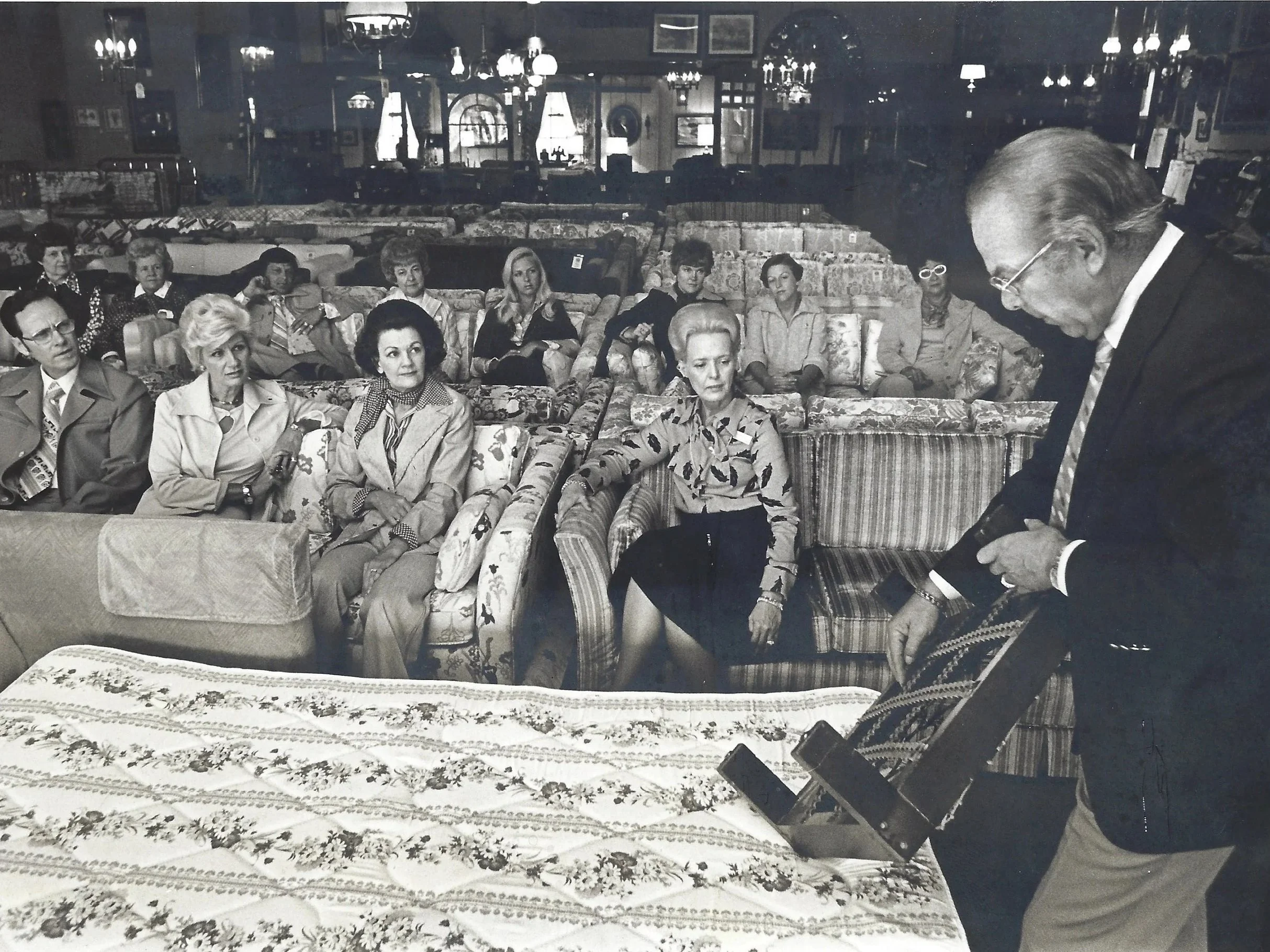 A man in a suit and glasses is demonstrating a device to a group of women sitting on sofas, in a store filled with furniture.