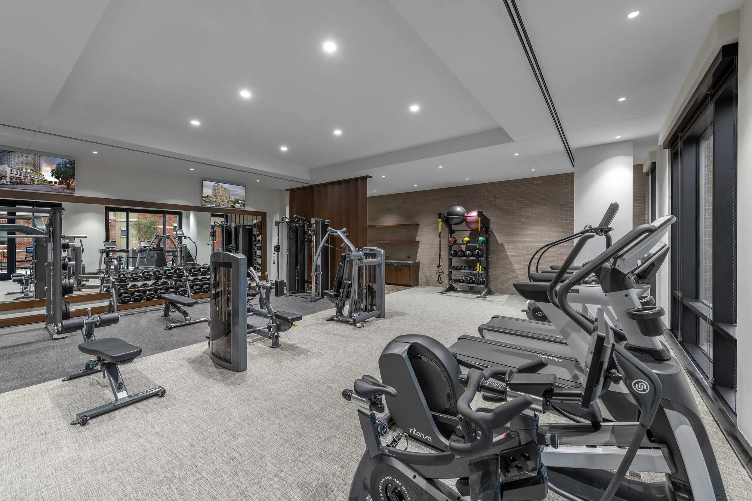 Well-lit gym with exercise equipment including treadmills, ellipticals, weight racks, and benches, with large windows and wall-mounted TVs.