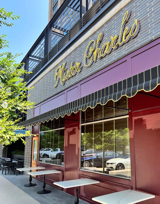 Exterior of a restaurant named Master Charles with large glass windows, a purple facade, and a black-and-yellow striped awning, with outdoor seating and parked cars reflected in the windows.