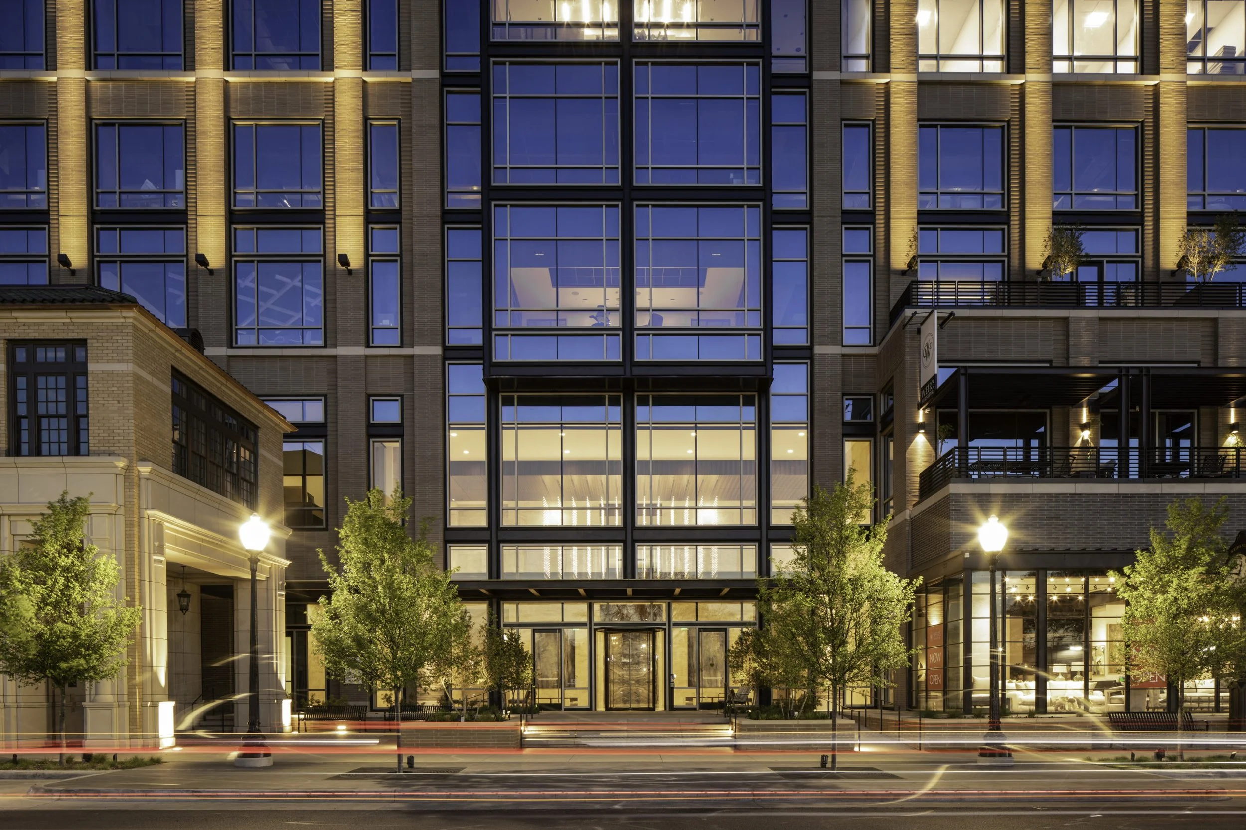 Modern glass-front building at dusk with illuminated interior, surrounded by small trees, streetlights, and blurred moving cars.