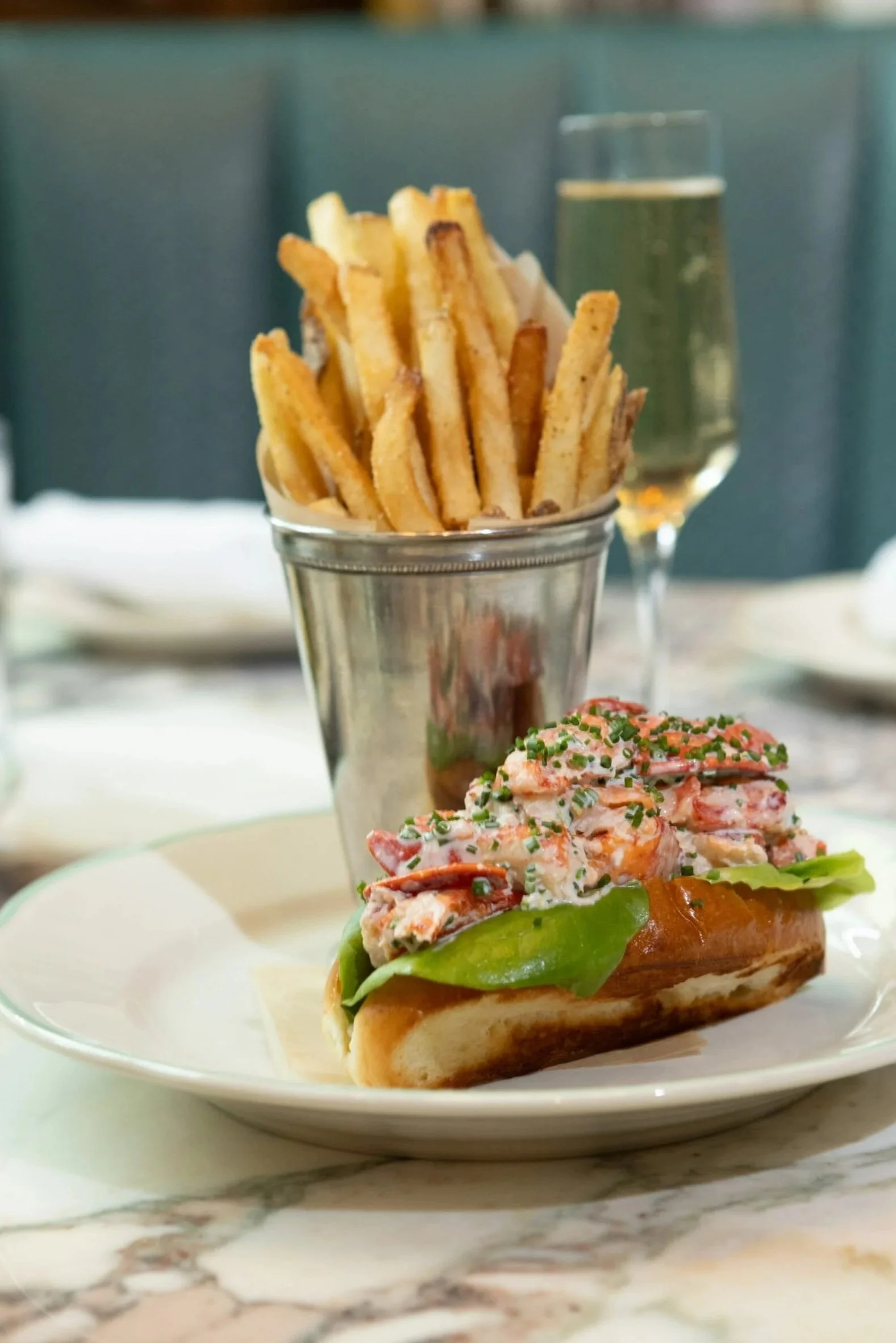 Lobster roll with lettuce on a bun, served on a white plate, with a side of French fries in a silver cup and a glass of champagne in the background, on a marble table.