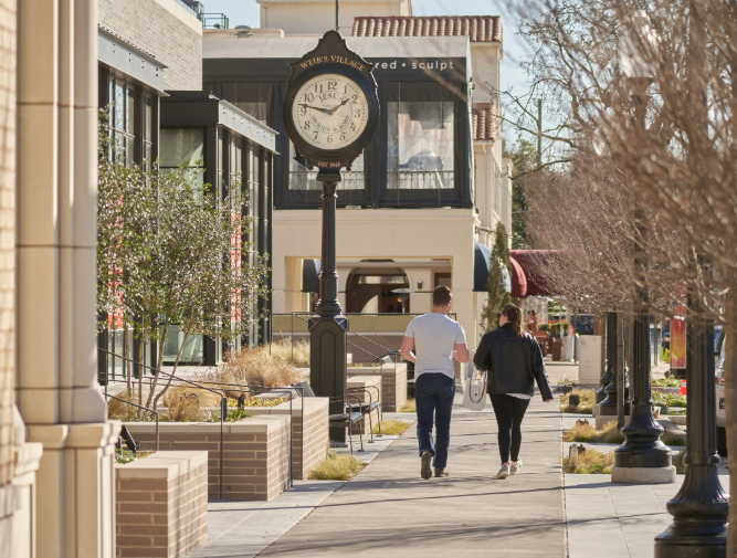 A man and a woman walking down a sidewalk in a shopping or downtown area with storefronts, trees, and a street clock showing 2:20.