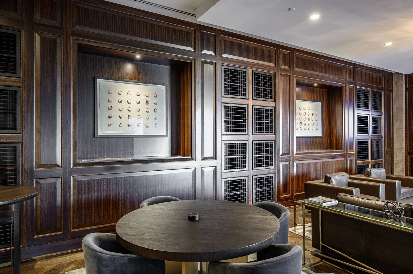 A luxurious room with dark wood paneling, framed artwork, a round table with chairs, and a leather sofa with a glass side table.