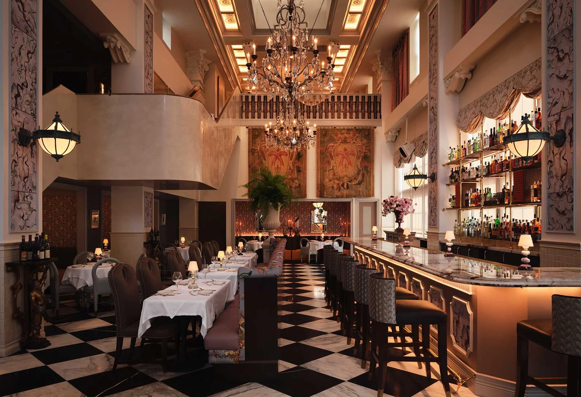 Elegant restaurant interior with checkered black and white marble floor, white tablecloth-covered tables, and a bar area with backlit shelves of liquor bottles. Crystal chandeliers hang from the ornate ceiling, and large windows with decorative curtains provide natural light. Artwork and floral arrangements adorn the space, creating a luxurious atmosphere.