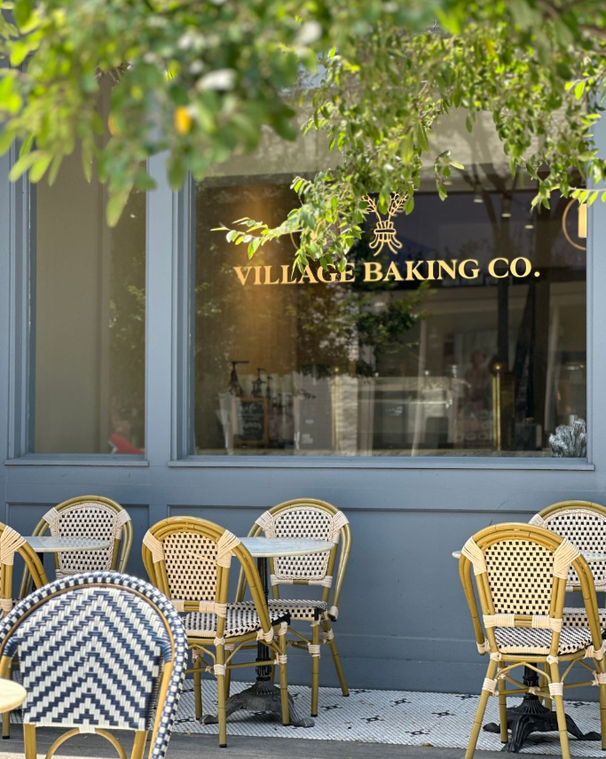 Empty outdoor seating area with patterned chairs outside Village Baking Co. store, with green tree branches overhead.