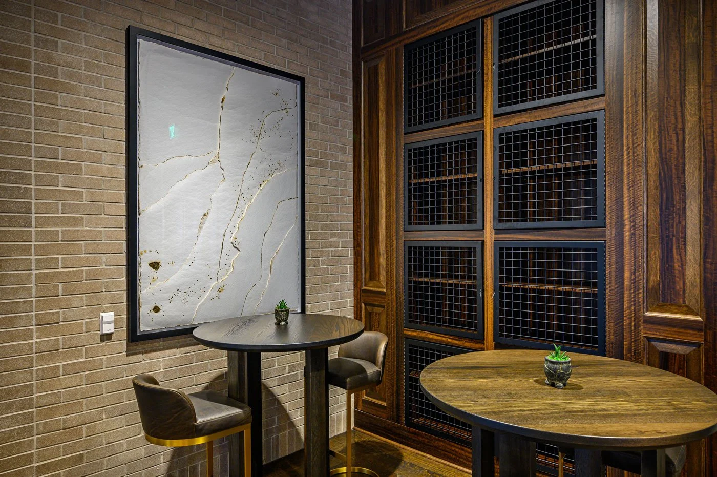 Interior of a room with brick and wooden walls, featuring two round tables with small potted plants, one with two chairs and the other empty, and a large abstract white and gold artwork on the brick wall.