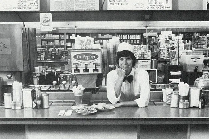 A woman working at a soda fountain counter in a classic diner, with various beverages, syrups, and ice cream supplies behind her.