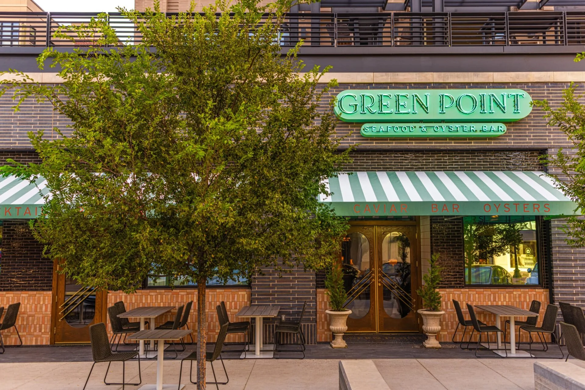 Exterior of Green Point Seafood & Oyster Bar with outdoor seating, a striped green and white awning, a large tree, and potted plants.