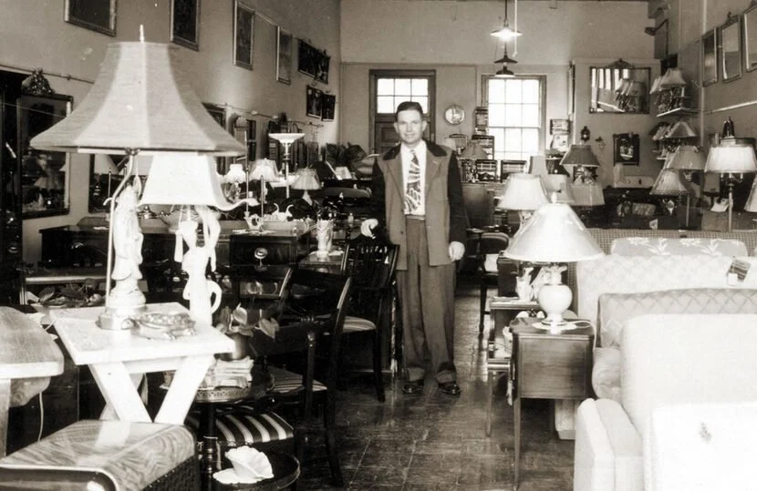 A man in a suit standing inside a vintage furniture store with various lamps, tables, and chairs on display.