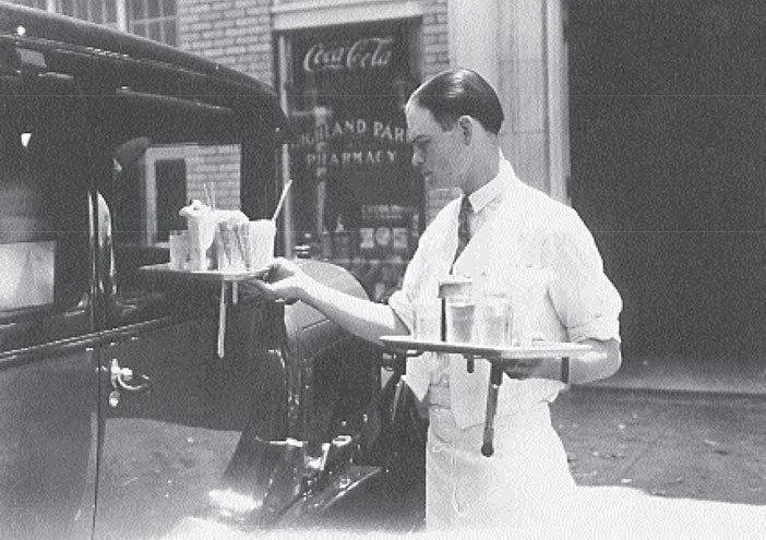 A man dressed in vintage attire, likely from the early 20th century, serving drinks from a tray into a car window in front of a Coca-Cola sign.