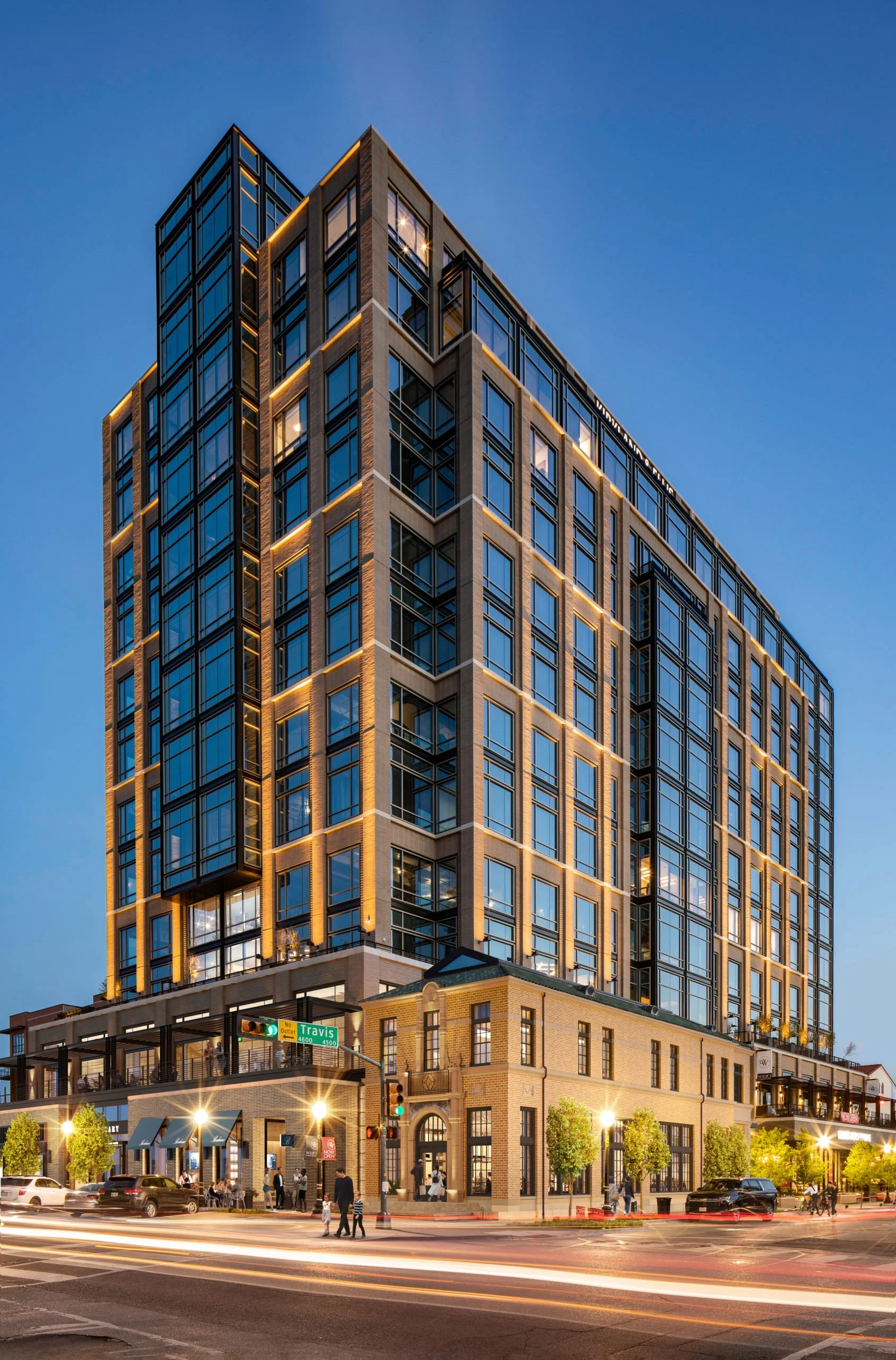 A tall modern building with large glass windows and exterior lighting, located at a street corner with people walking and cars driving by at dusk.