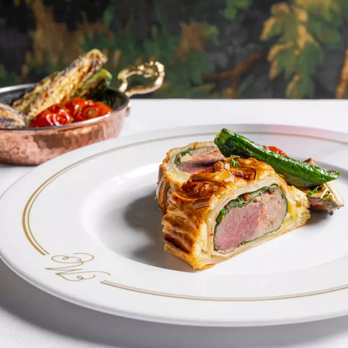 A plated dish of Beef Wellington, with sliced meat, pastry crust, and green vegetables, on a white plate with gold trim, with a copper dish of roasted vegetables in the background.