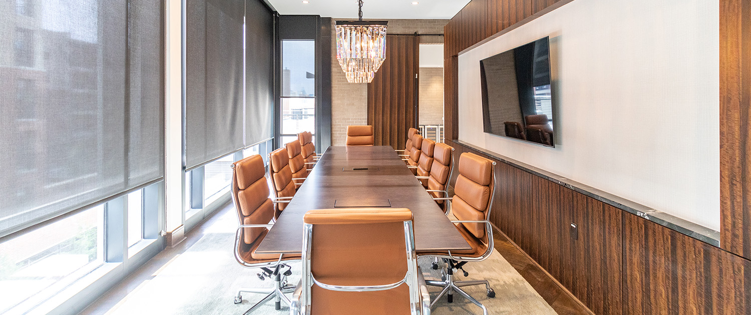 Conference room with a long wooden table surrounded by tan leather chairs, large windows with blinds, a flat-screen TV on the wall, and a chandelier hanging from the ceiling.