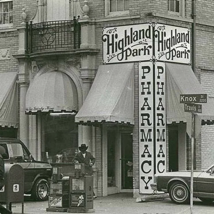 Vintage pharmacy storefront labeled 'Highland Park Pharmacy' at the corner of Knox and Travis streets, with a man in a suit and hat standing outside by a public phone booth, and two parked cars nearby.