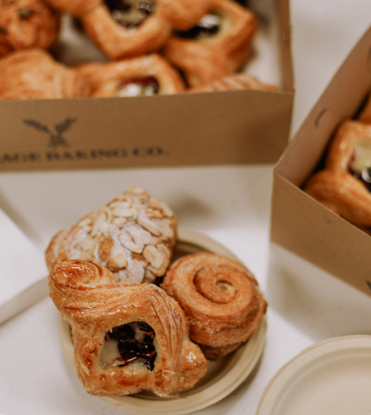 Various pastries including an almond croissant, a cinnamon roll, and a Danish pastry with fruit filling on a plate with boxes of more pastries in the background.