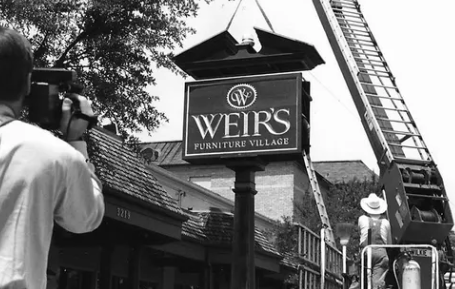 Photographer taking a photo of a sign for WEIR'S Furniture Village outside a building.