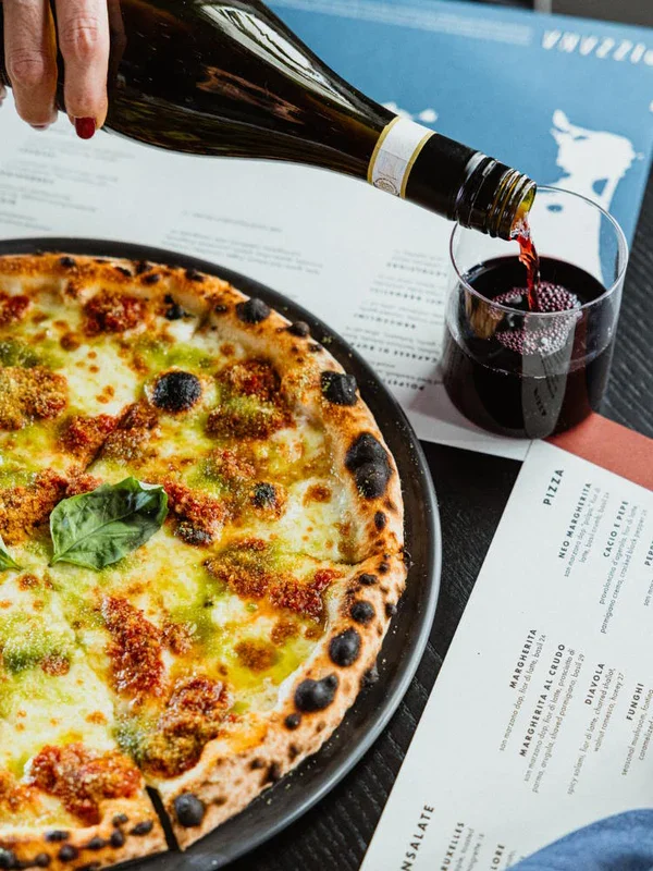 A hand pouring red wine into a glass beside a cheese pizza with basil leaves on top, on a table with a menu.