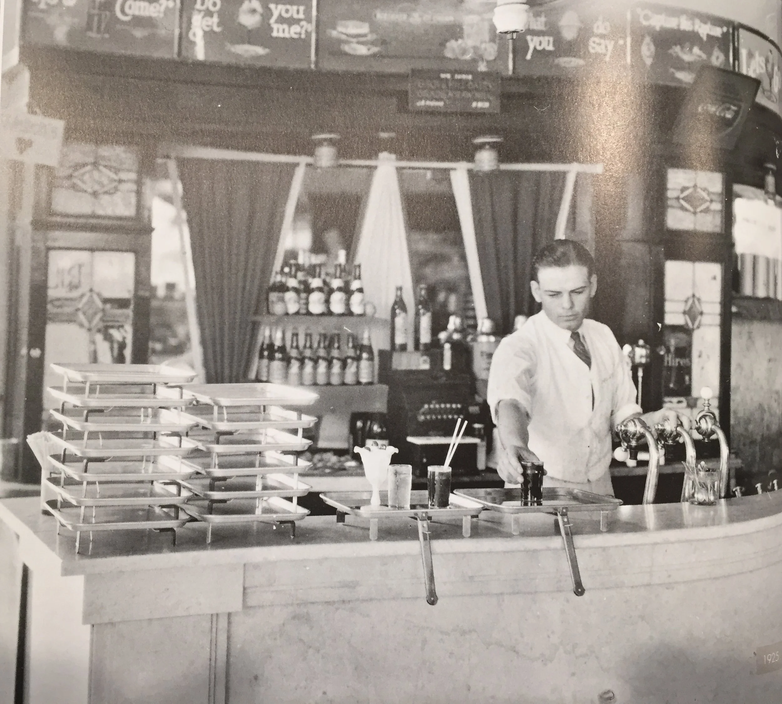 A man in a white shirt and tie behind a bar counter with empty trays, bottles, and glasses, preparing drinks in a vintage bar or cafe setting, with shelves of bottles and signs in the background.