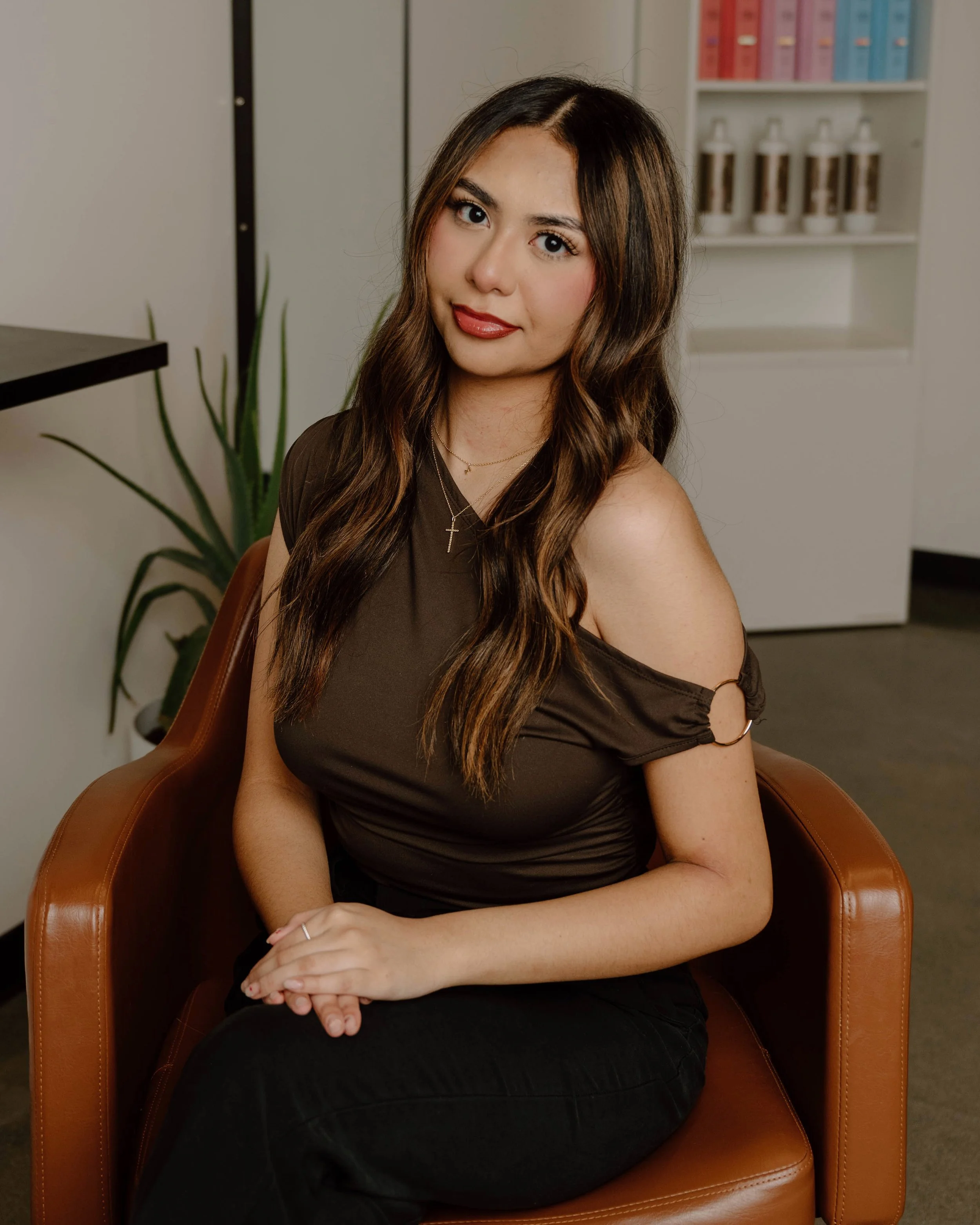 Young woman with long wavy brown hair wearing a dark brown off-shoulder top sitting in a brown leather chair in an office setting, with a plant and white cabinets with bottles in the background.