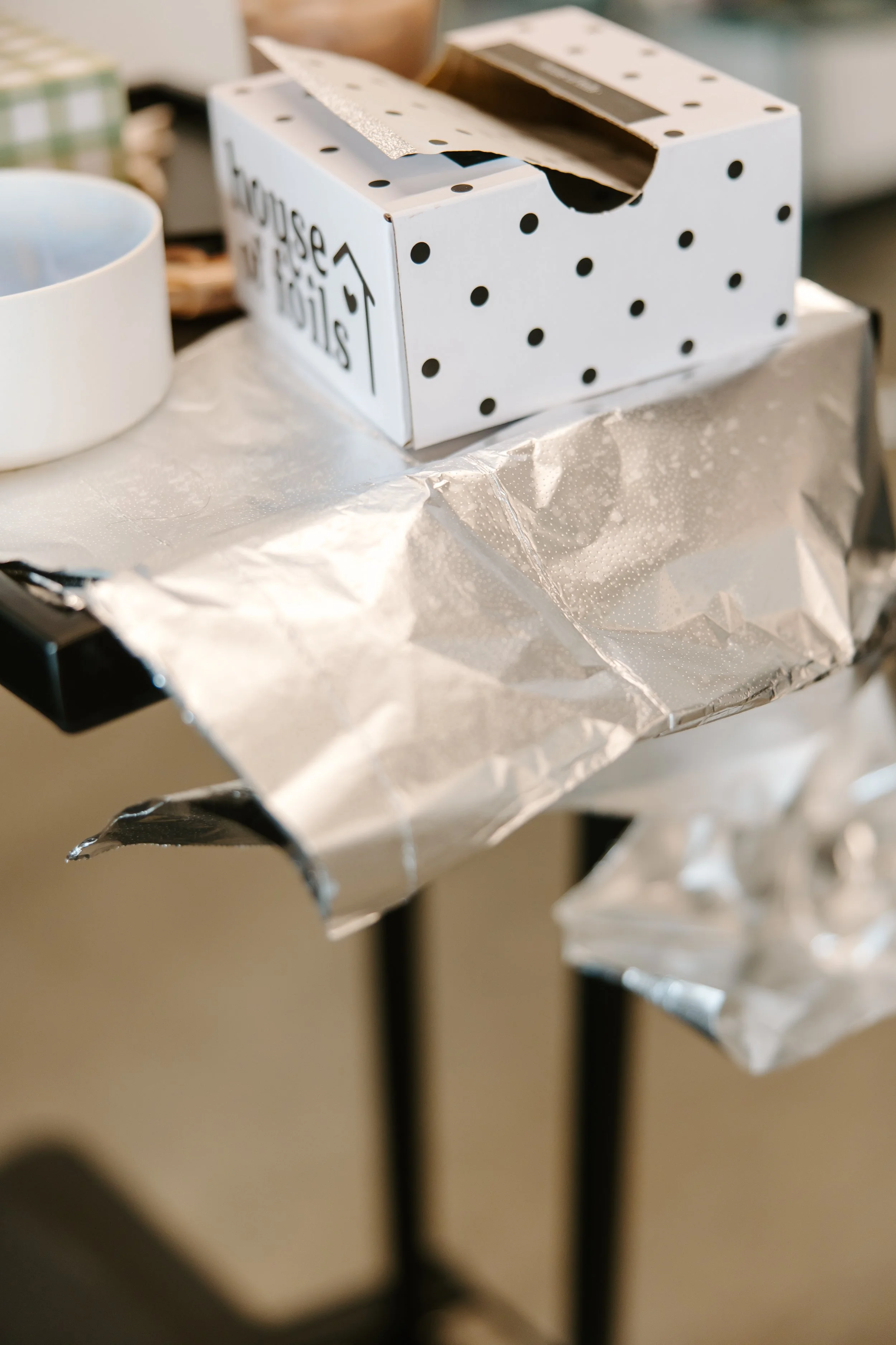 White tissue box with black polka dots and a 'Home Sweet Home' sign on the side, placed on a table with crinkled silver wrapping paper.