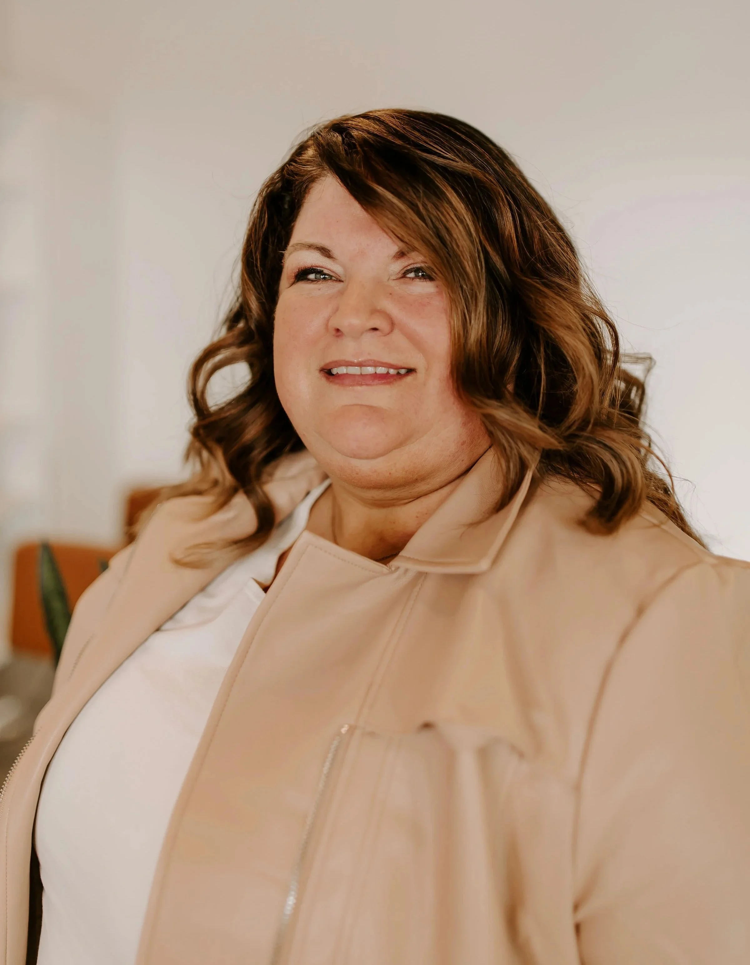 A woman with brown, wavy hair smiling, wearing a beige jacket and white top, in a room with light-colored walls.