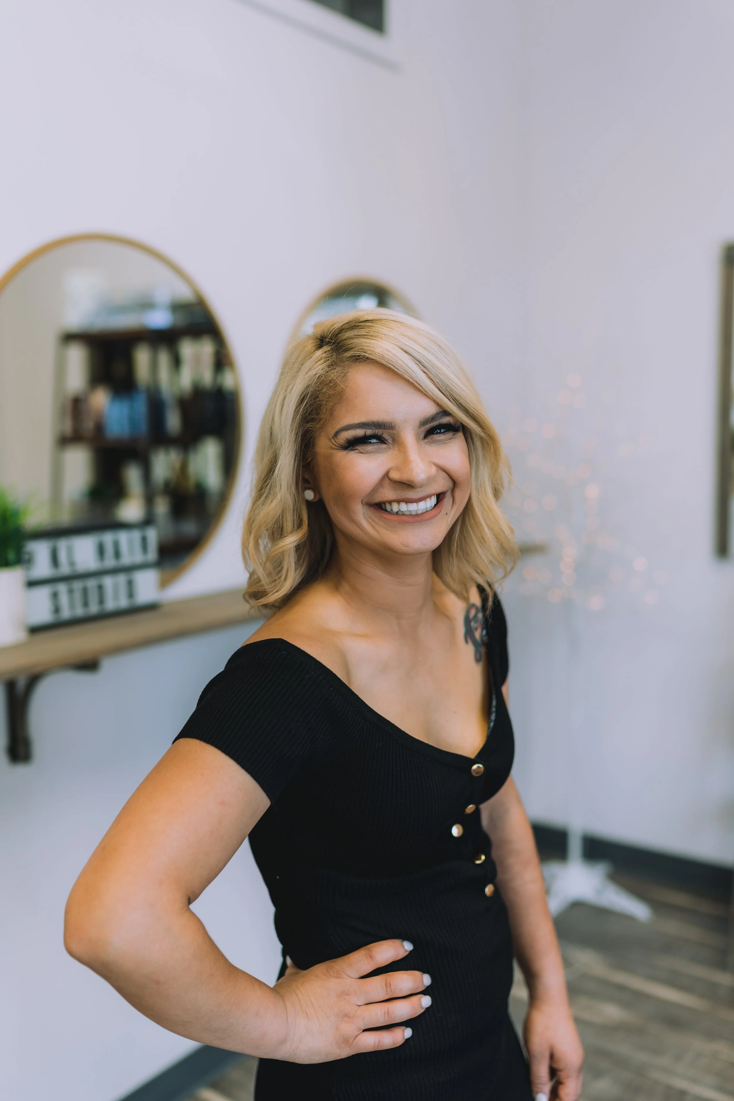 A woman with blonde hair smiling and posing indoors, wearing a black top with buttons and a tattoo on her shoulder.