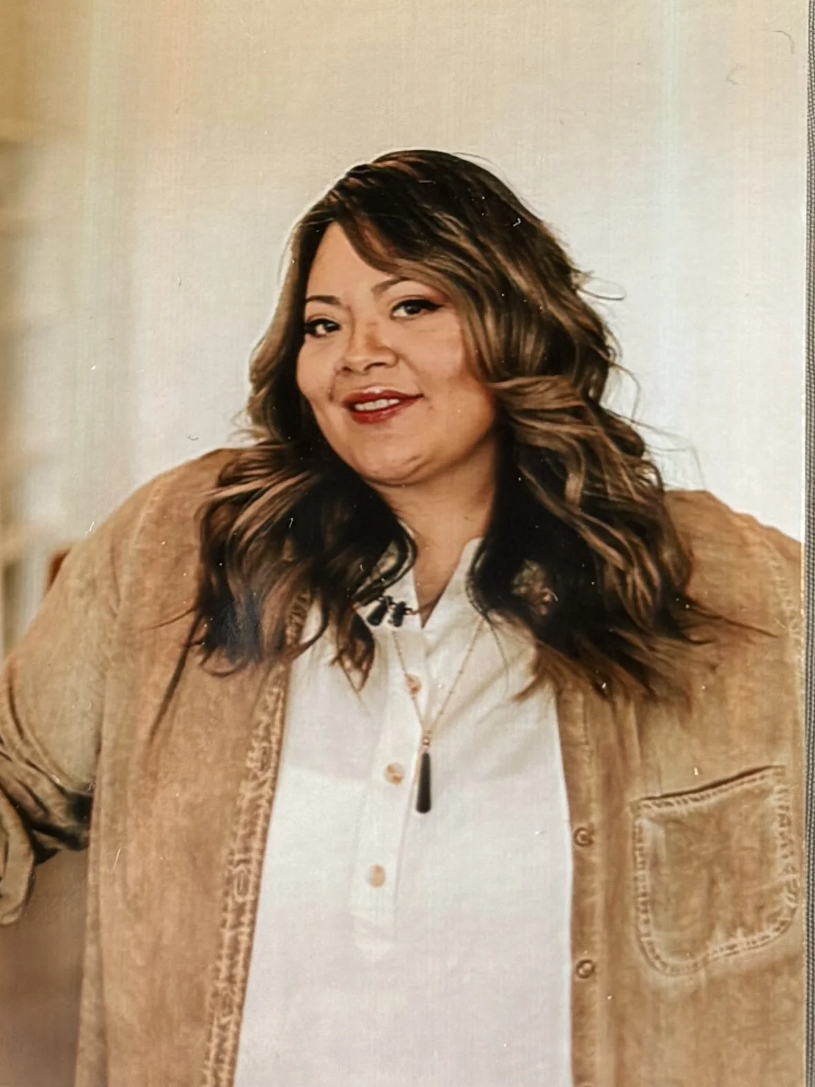 A woman with shoulder-length wavy hair, wearing a white top and a light brown jacket, standing indoors with a warm smile.