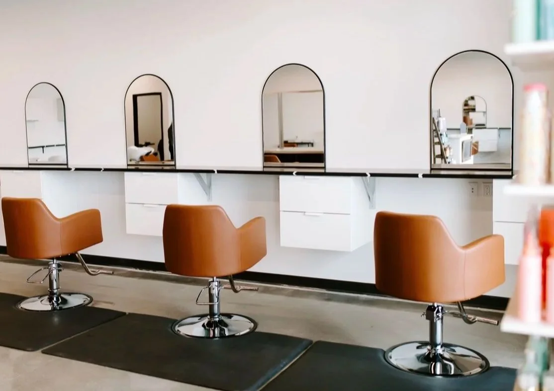 Salon chairs in front of mirrors at a hair salon, with white walls and shelves.