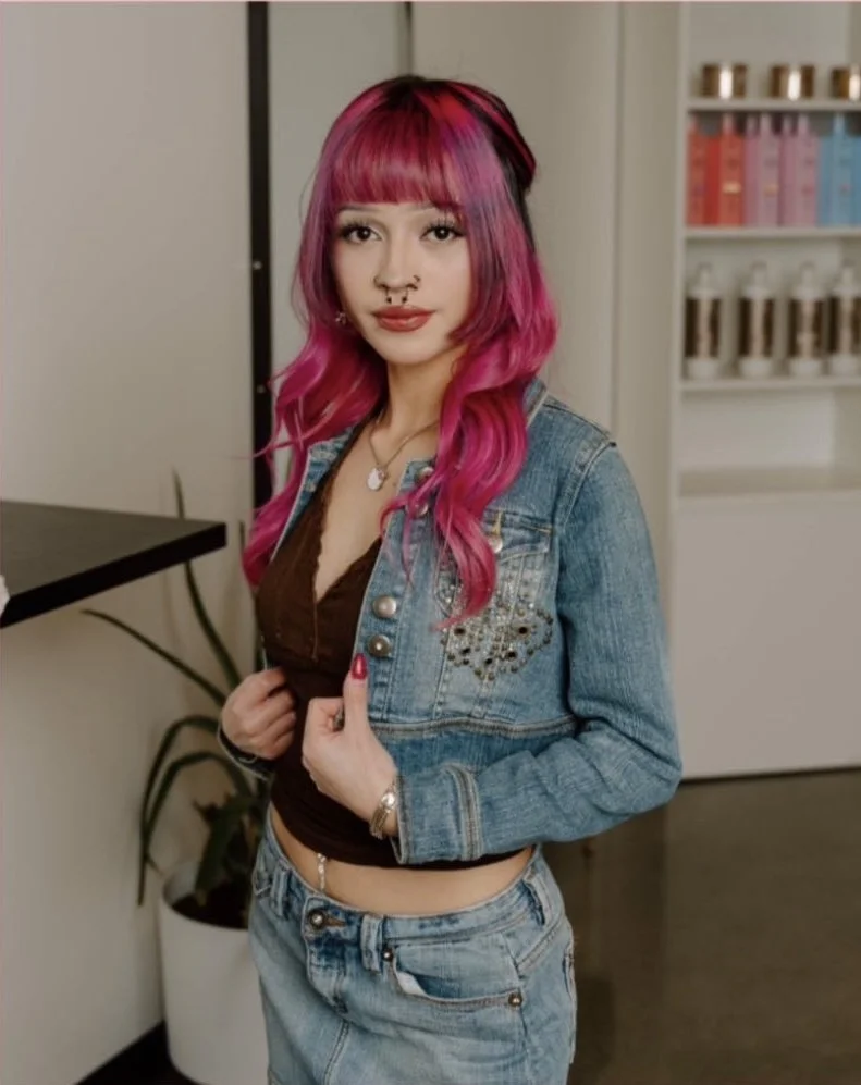 Young woman with pink hair, wearing a denim jacket and jeans, standing indoors with shelves of bottles behind her.
