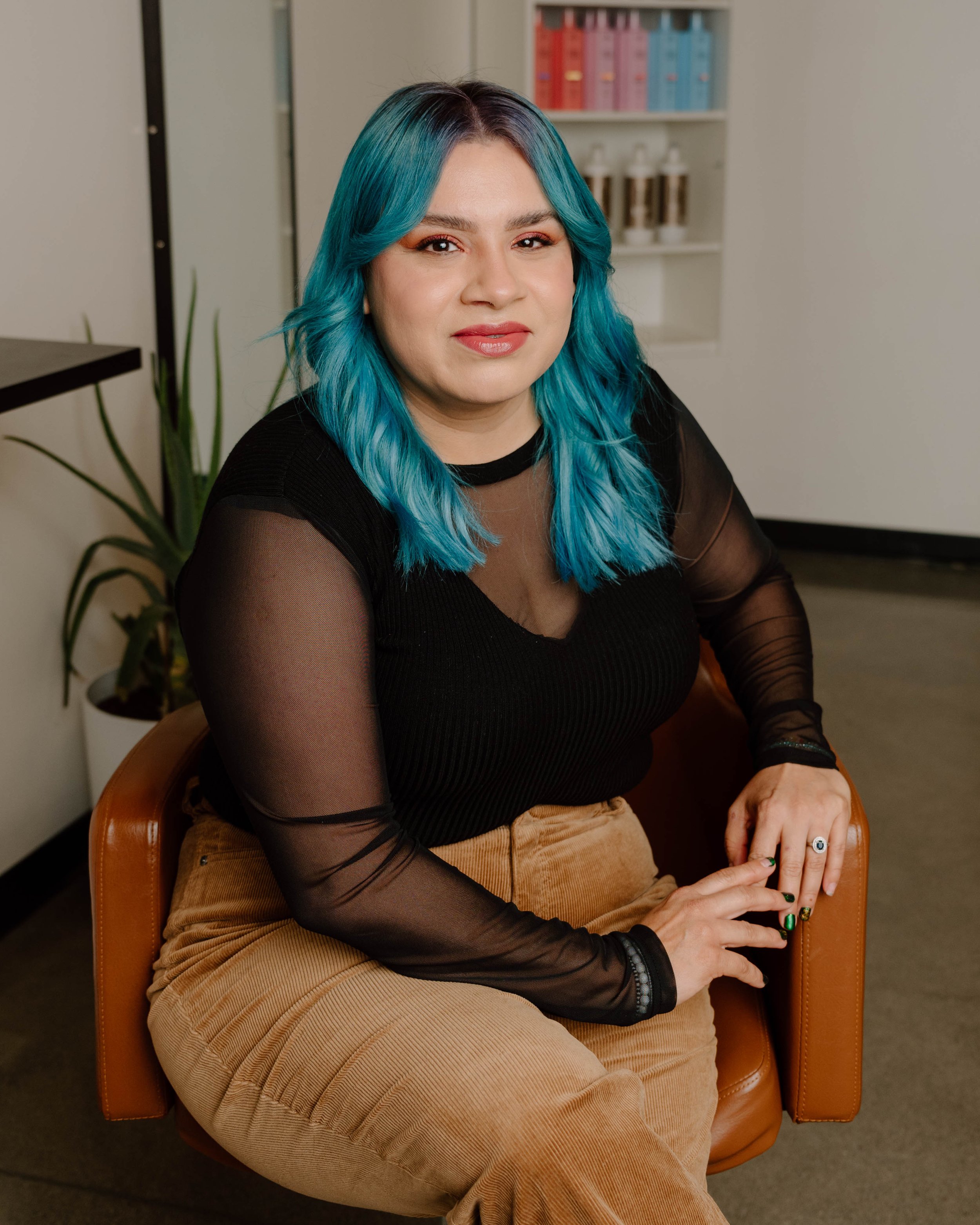 Woman with vibrant blue hair sitting on a brown chair, wearing a black sheer long-sleeve top and beige pants, in an office or studio setting.